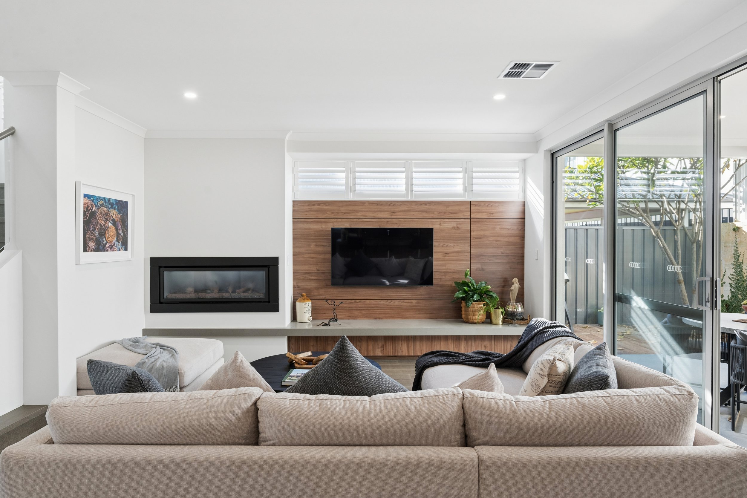 Living room with beige sofa, gray pillows, wall-mounted TV on wooden accent wall, fireplace below TV, plants on side, large glass sliding door opening to outdoor patio with trees.
