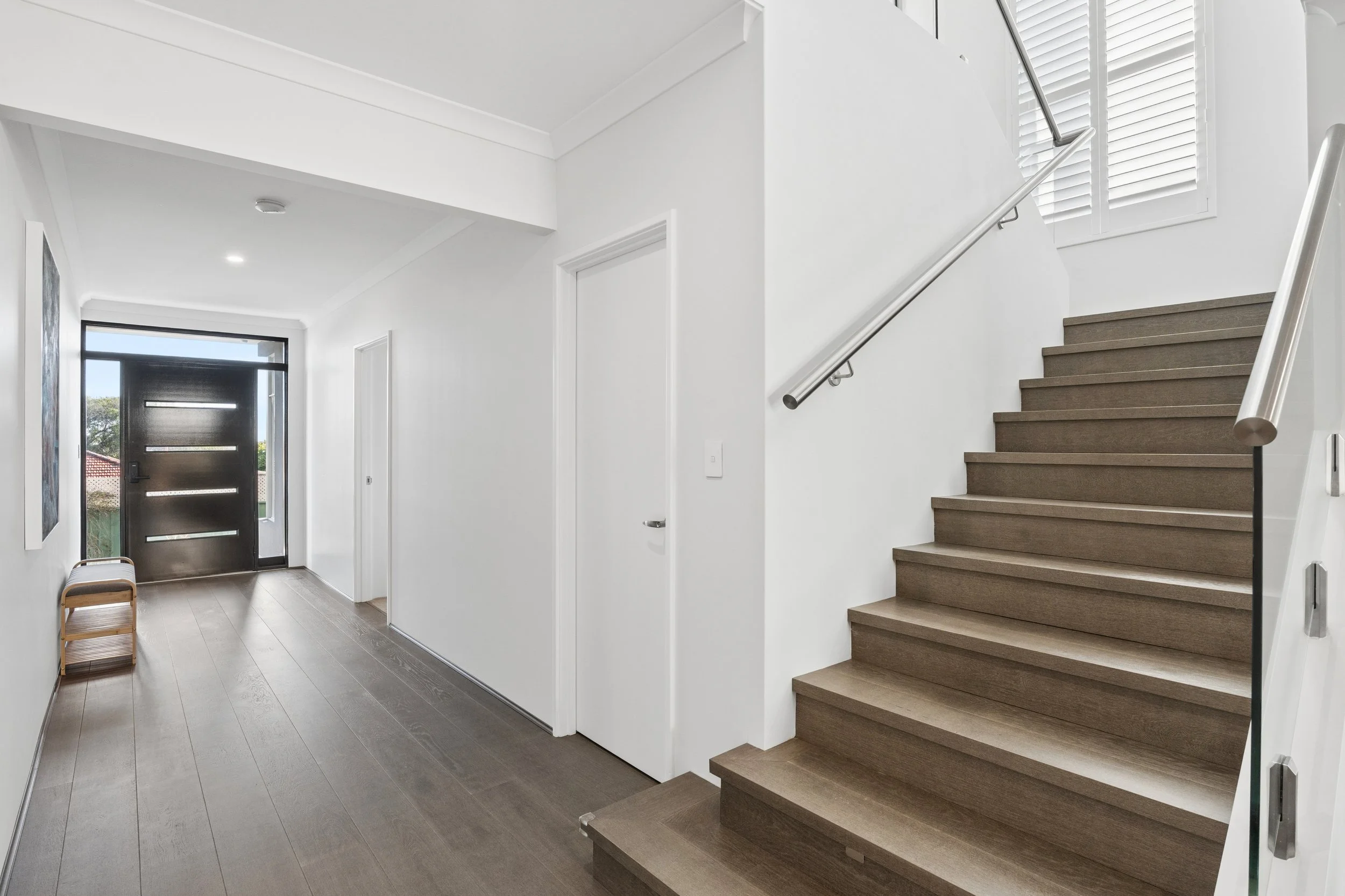 Modern interior entryway with dark front door, white walls, staircase with dark wood steps, window shutters, and flooring, minimal decor.