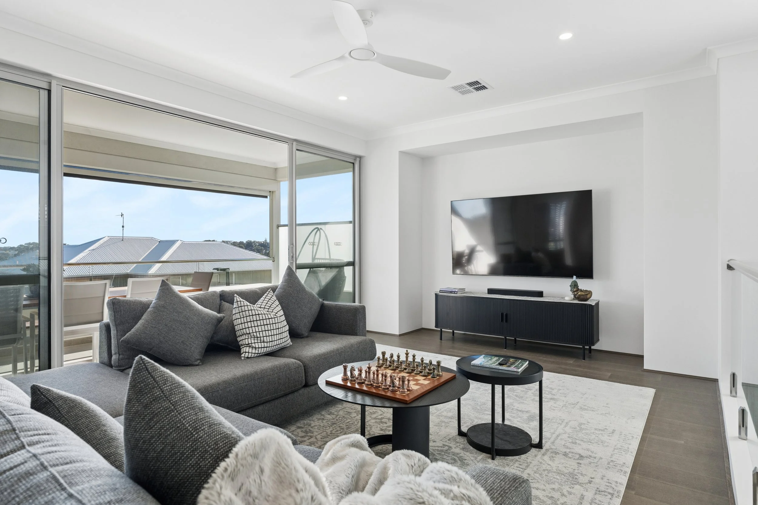 Modern living room with large open sliding glass doors leading to a balcony, featuring a gray sectional sofa with pillows, a chess set on a coffee table, a black TV on a stand, and a ceiling fan, with a view of rooftops through the windows.