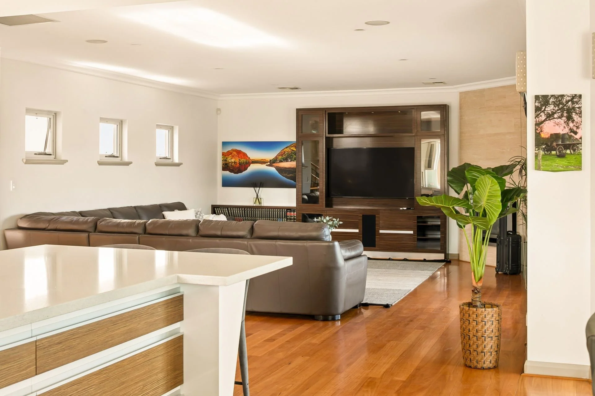 Living room with brown leather sectional sofa, wall-mounted flat-screen TV, large potted plant, three small windows, and a painting of a landscape with water and hills.