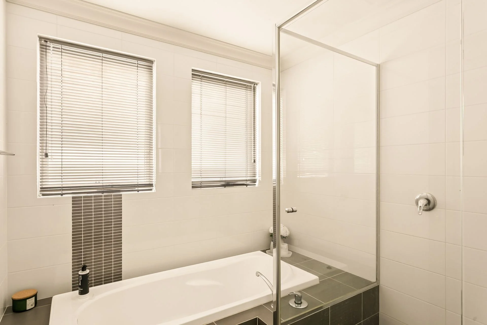 Modern bathroom with a bathtub and a walk-in shower, two windows with blinds, white tiled walls, and gray tile floor.