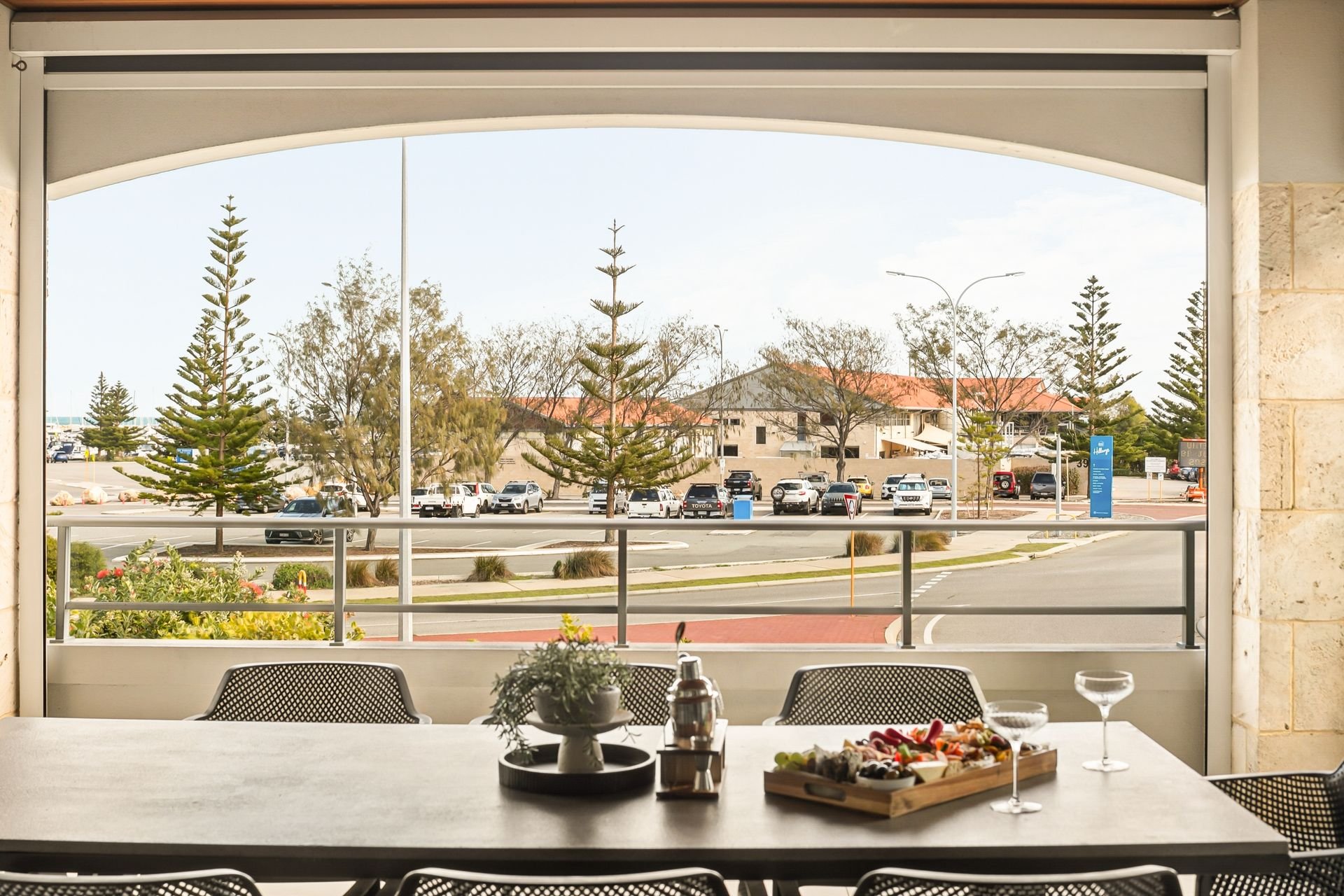 Indoor dining area with a table set with a cheese platter, wine glass, and potted plant, overlooking an outdoor parking lot with trees, buildings, and parked cars through large open windows.
