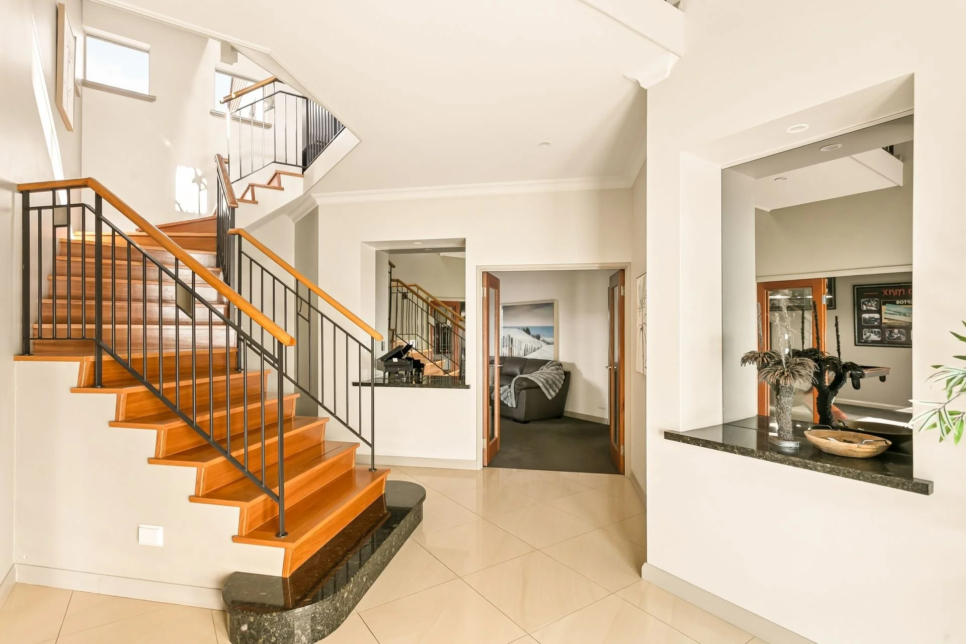 Interior of a modern home with wooden staircase, black metal railings, white walls, and an open living space including a black sofa, a grand piano, and decorative plants.