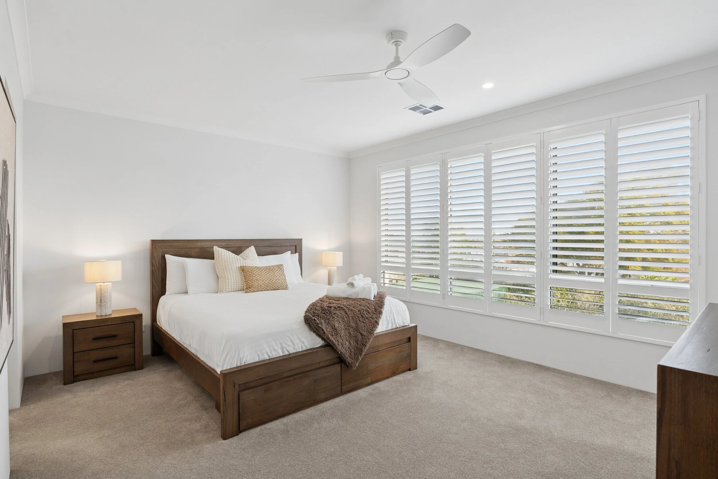 Bedroom with a wooden bed frame, white bedding, and a brown blanket. Two bedside tables with lamps. Large window with white plantation shutters, carpeted floor, white walls, and a ceiling fan.