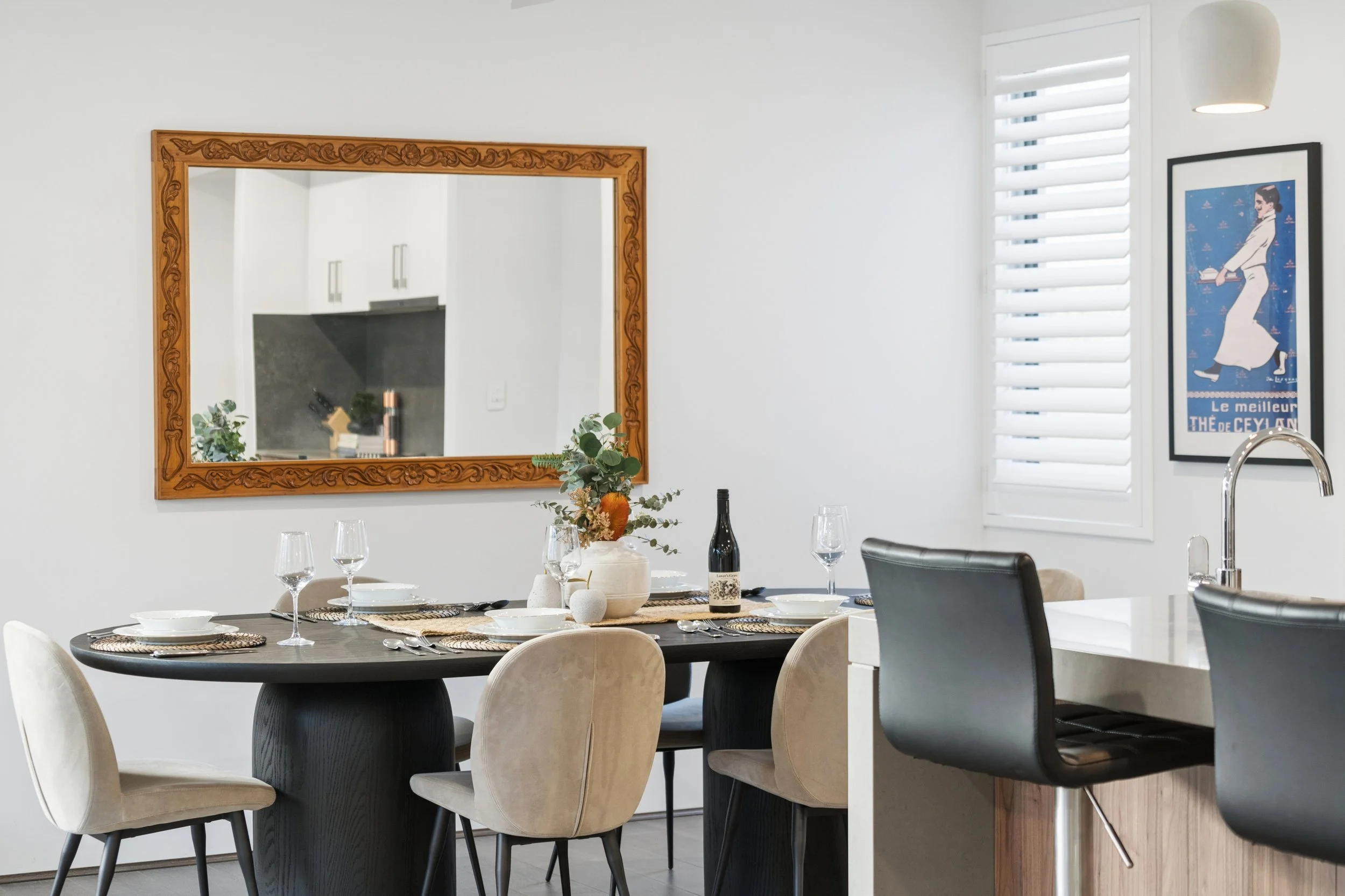 A modern dining room with a round black table set for a meal, surrounded by cream and black chairs. The wall features a large ornate wooden mirror and a framed vintage-style poster. A window with white blinds and a white pendant light are visible.