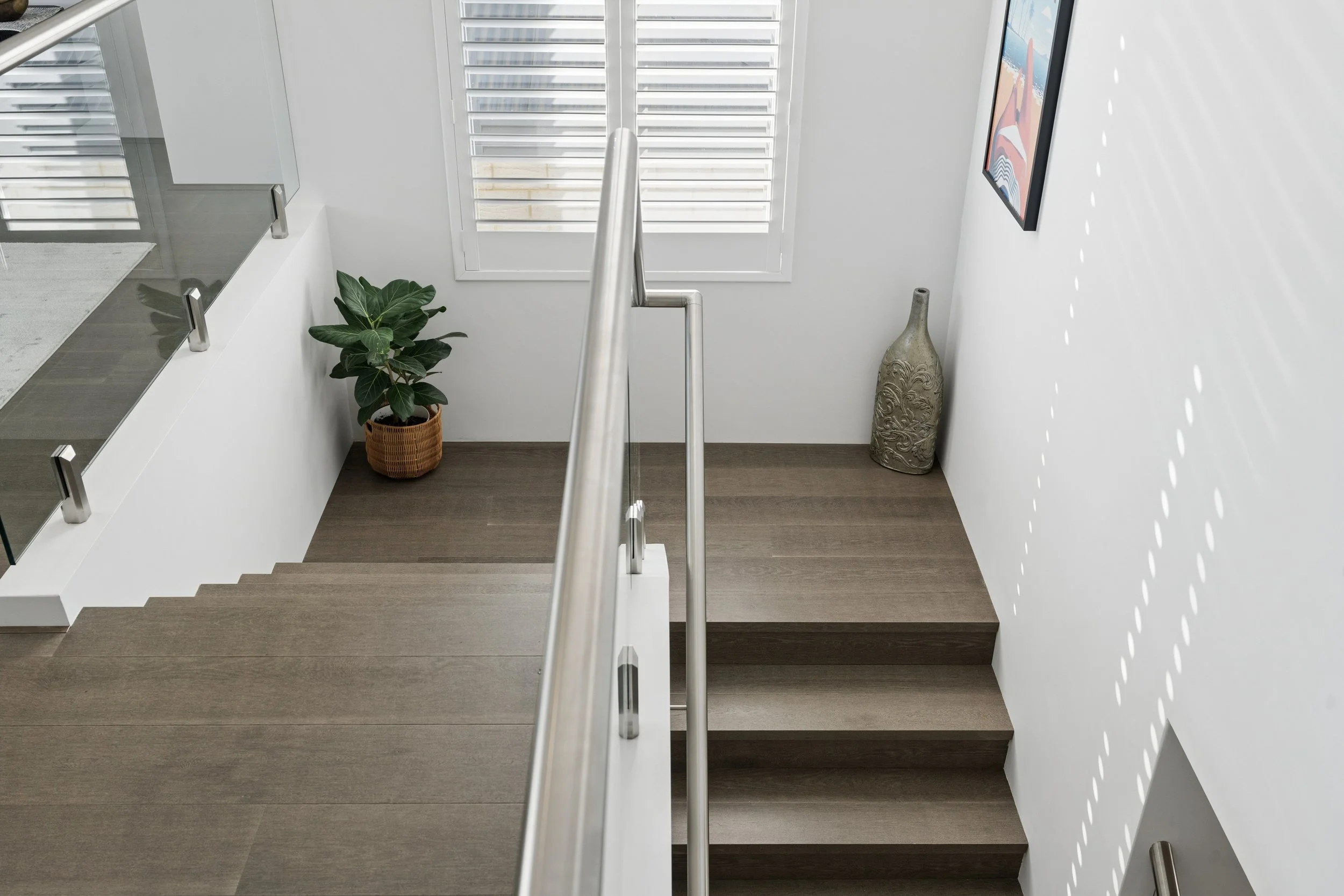 A staircase seen from the top with wooden steps, a middle stainless steel handrail, white walls, a recycling bag, a plant, and an art piece on the wall near a window with white shutters.