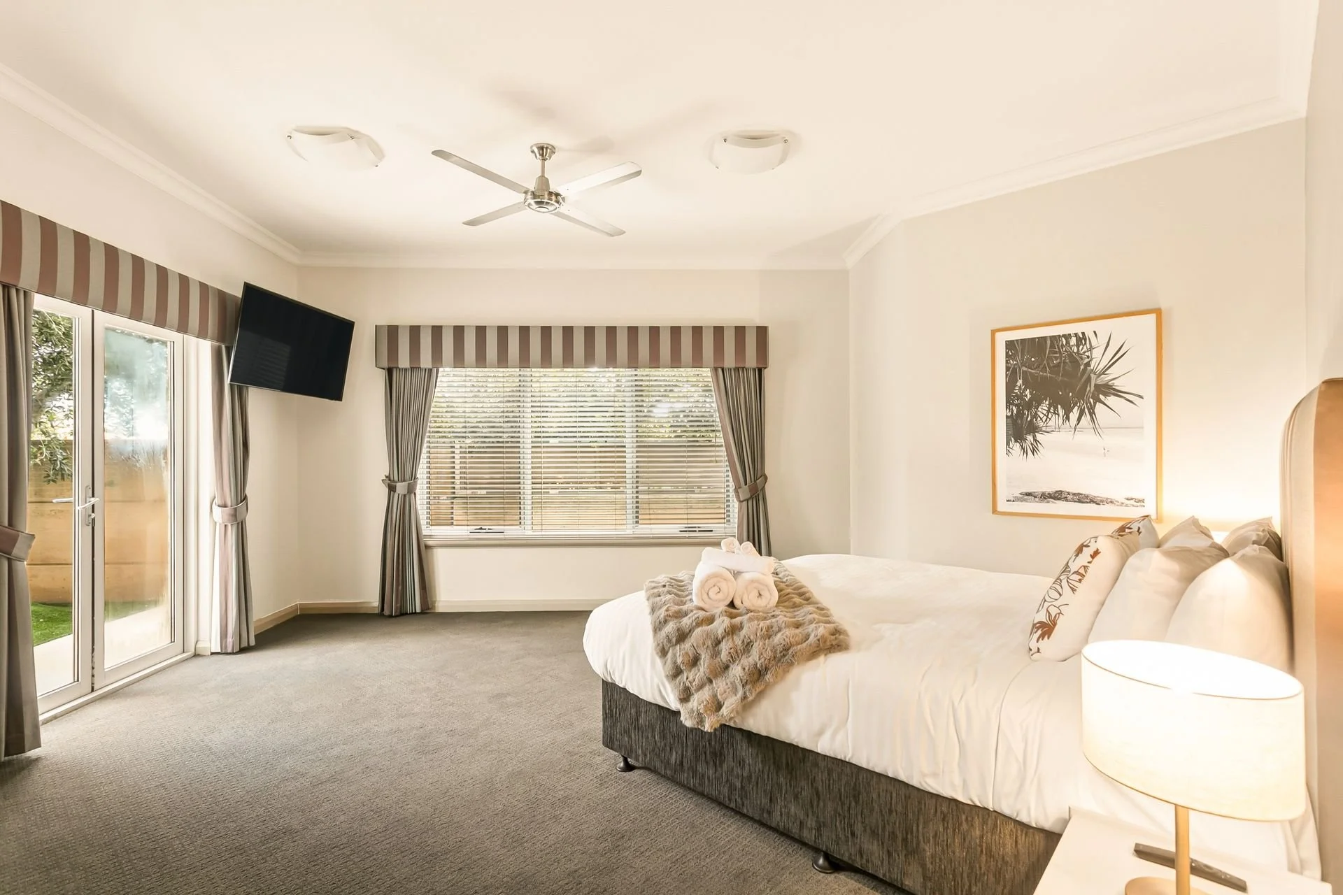 A cozy bedroom with a large bed covered in white linens, pillows, and a fur throw. A stack of rolled towels sits at the foot of the bed. There's a window with striped curtains and blinds, and a wall art featuring a tropical plant. The room has a ceil