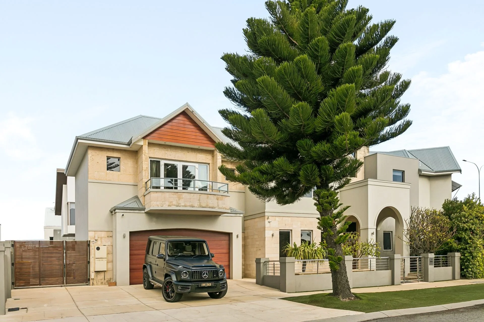 Modern house with a gray Mercedes-Benz G-Class SUV parked in front, a tall green pine tree in the front yard, and a driveway and fence in a suburban neighborhood.