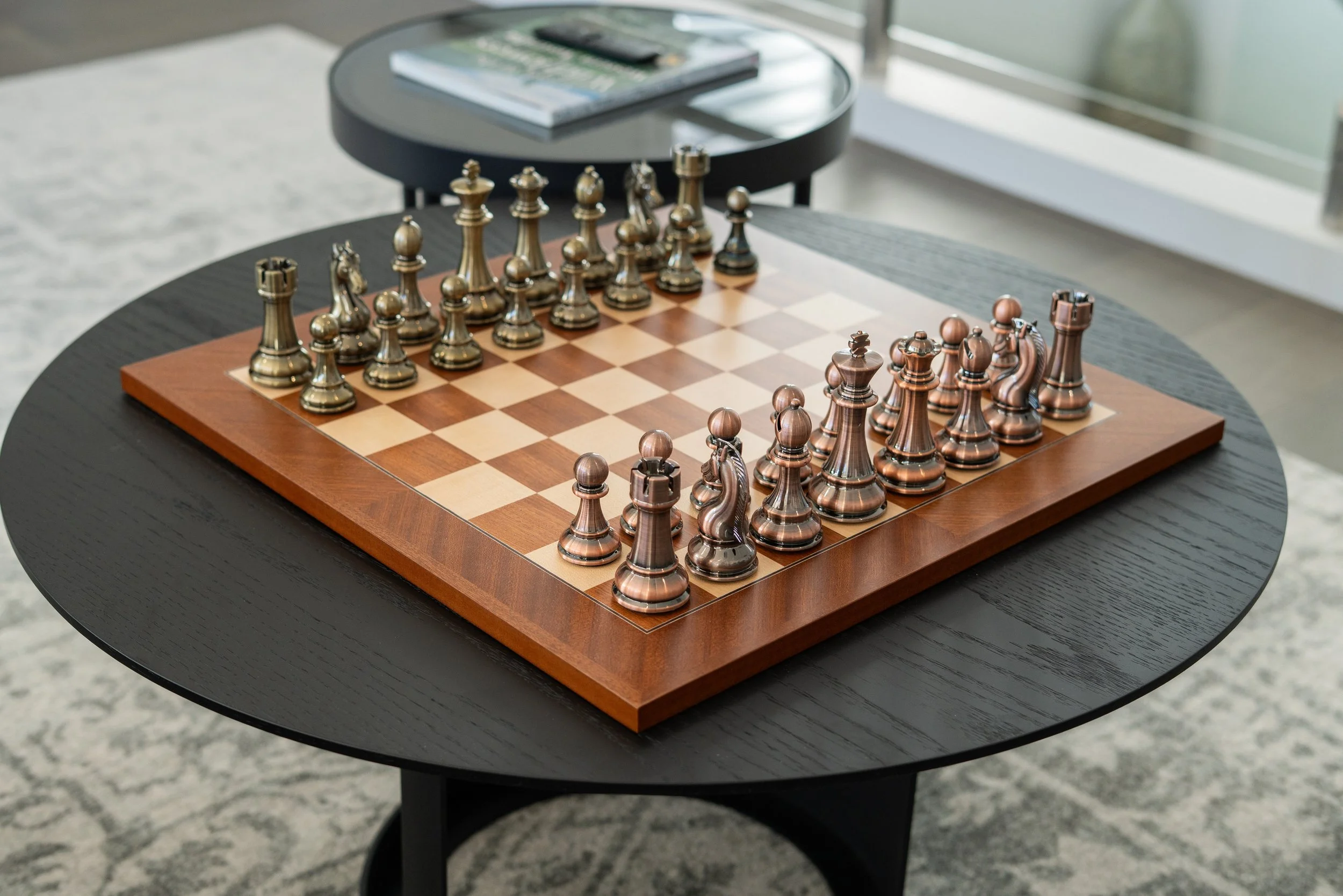 A chessboard with metallic chess pieces on a black round table, with a magazine and a remote control on a side table in the background.