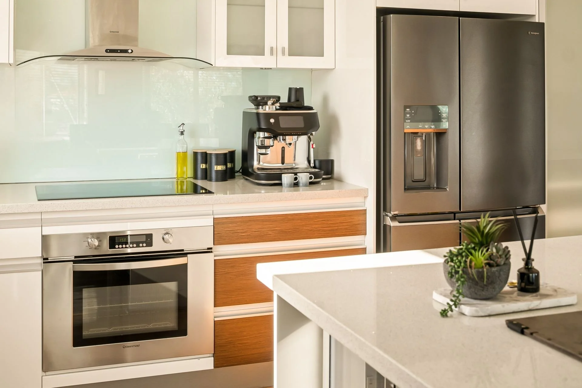 Modern kitchen with white and wood cabinets, stainless steel oven and refrigerator, coffee machine, black containers, and a small plant on a white countertop.
