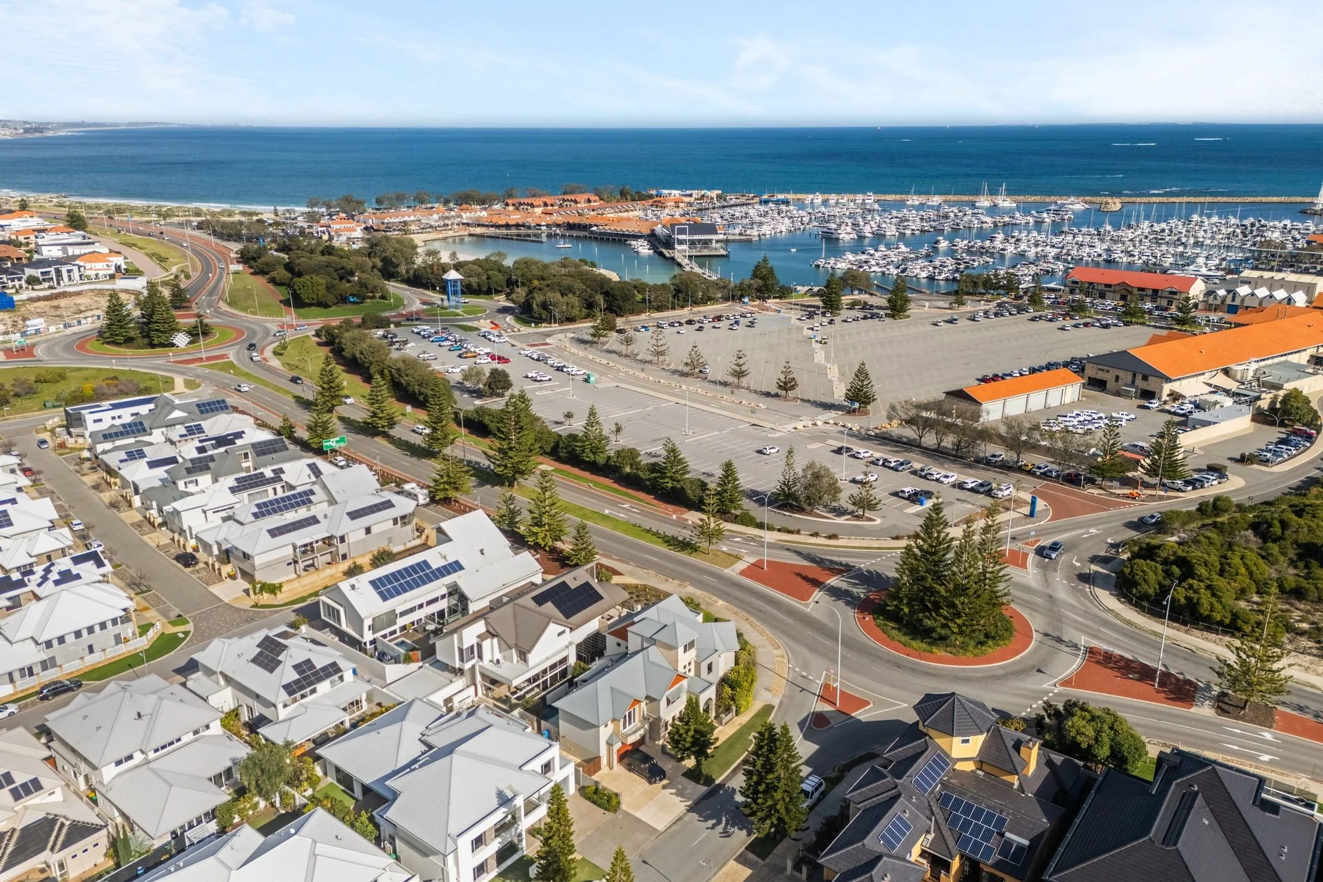 Aerial view of a coastal marina with numerous boats docked, surrounded by residential houses with solar panels and a parking lot, with the ocean in the background under a partly cloudy sky.