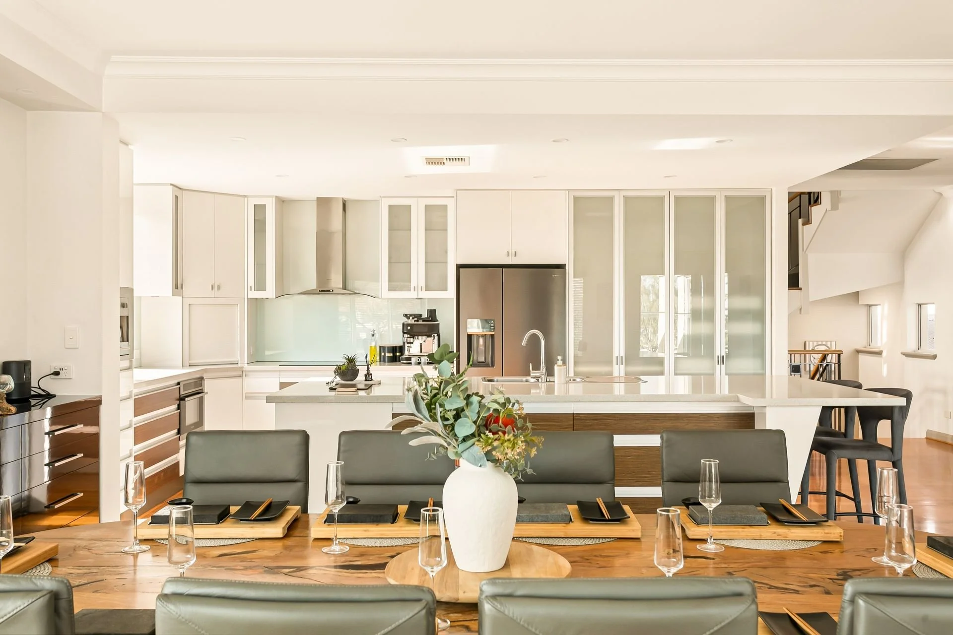 Modern, open-concept kitchen and dining area with a large kitchen island, white cabinetry, stainless steel refrigerator, and a dining table set with black dishes, glasses, and a white vase with flowers.
