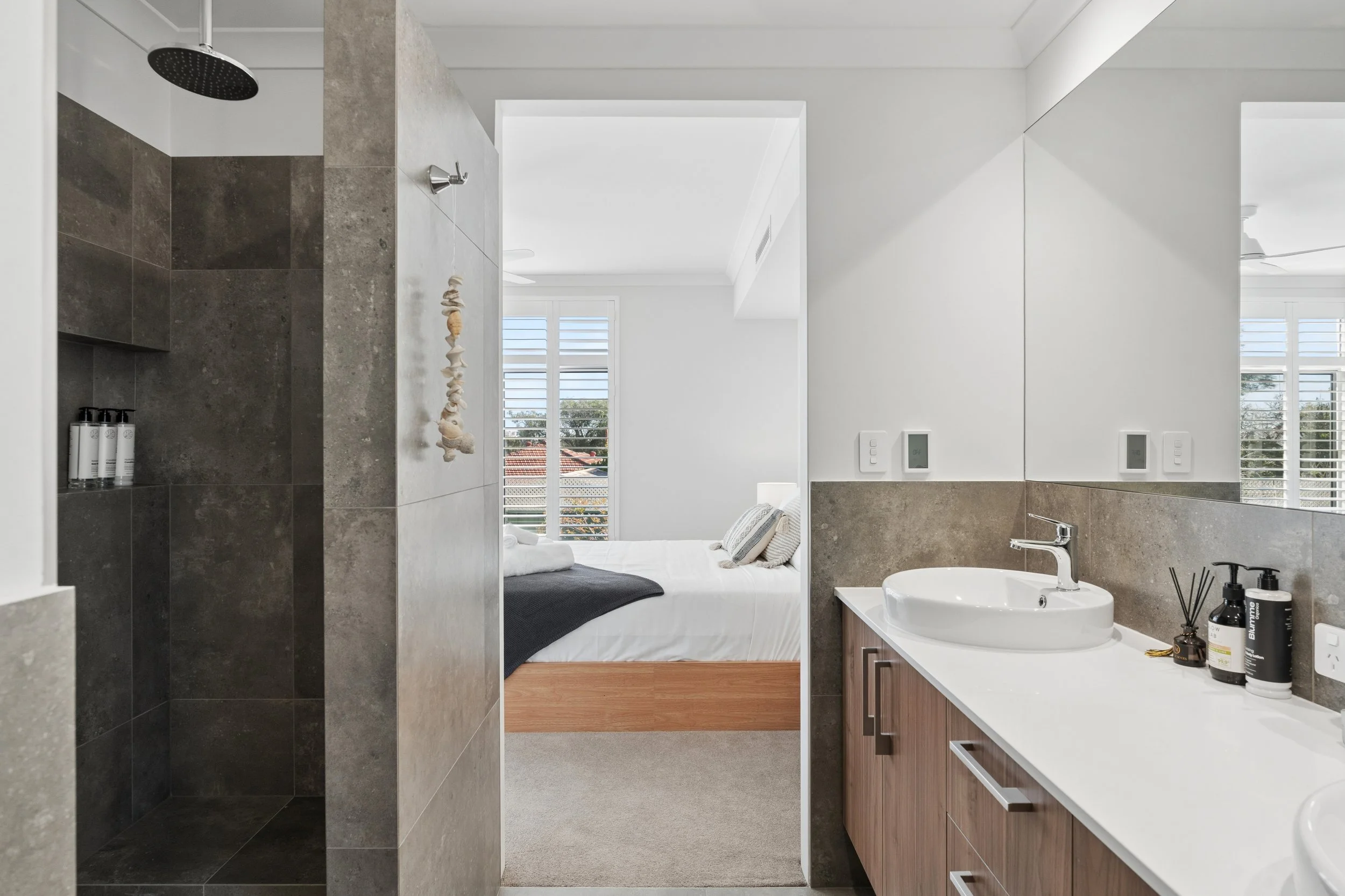 Modern bathroom with a view into a bedroom with white walls and a large window.