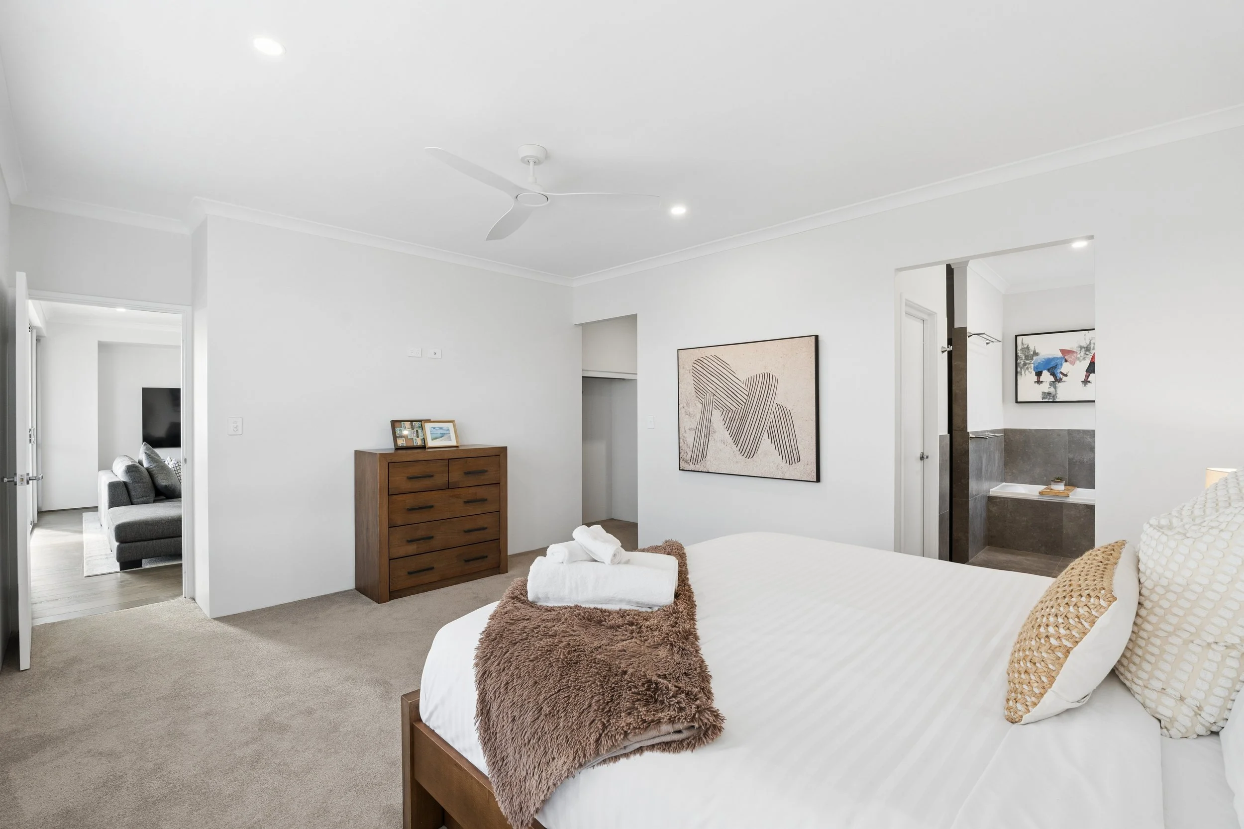 A bedroom with white walls and ceiling, featuring a wooden dresser, framed artwork, and a bed with white linens, pillows, and a brown furry throw. A bathroom with gray tiles and a framed picture is visible through an open doorway.