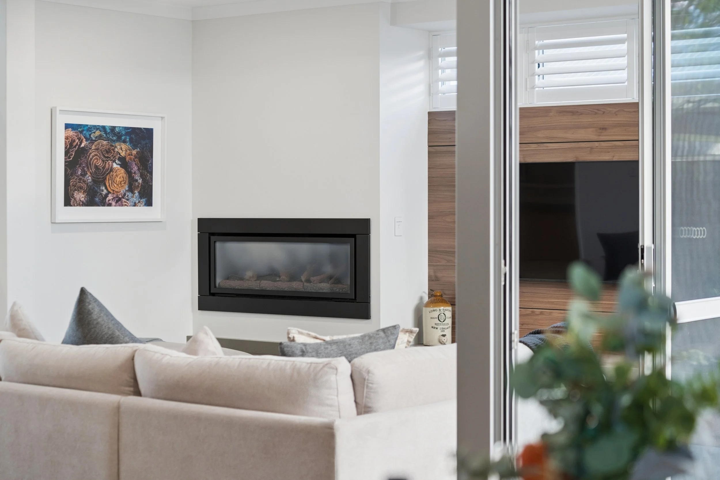 Living room interior with a beige sofa, a wall-mounted fireplace, wall art of corals, and large windows with white blinds.