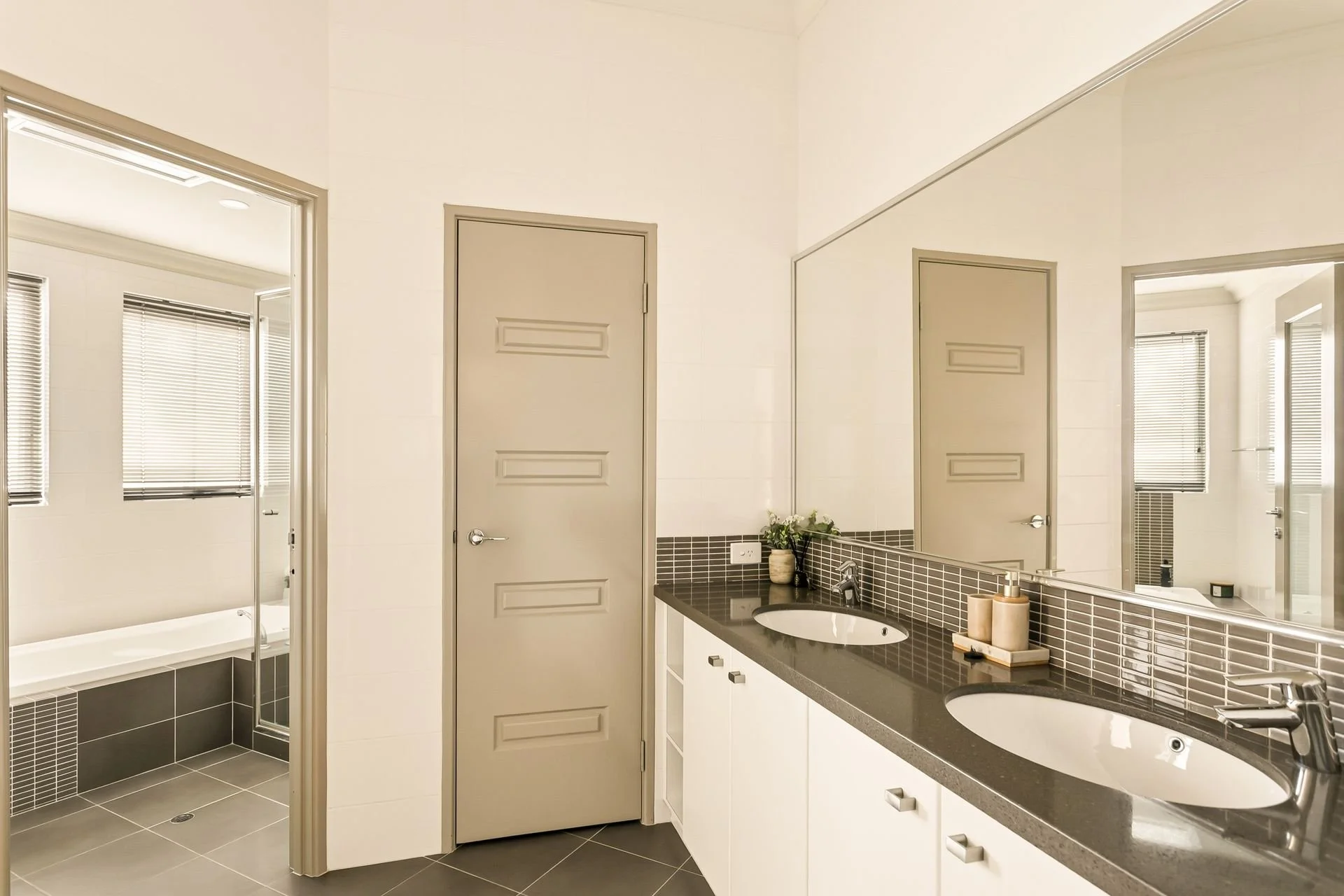 Modern bathroom with dual sinks, a large mirror, and beige doors. There is a vanity with a black countertop, plant, and bathroom accessories. A shower area with glass doors and blinds on the windows are visible.