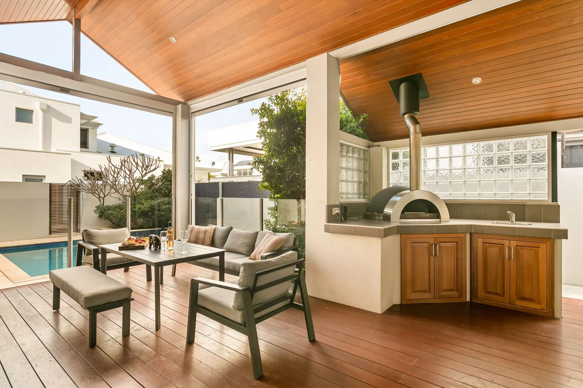 Outdoor patio area with cushions and a table near a pool, adjacent to a kitchen with a wood stove and cabinets, open to the outside with large glass windows and a wood ceiling.