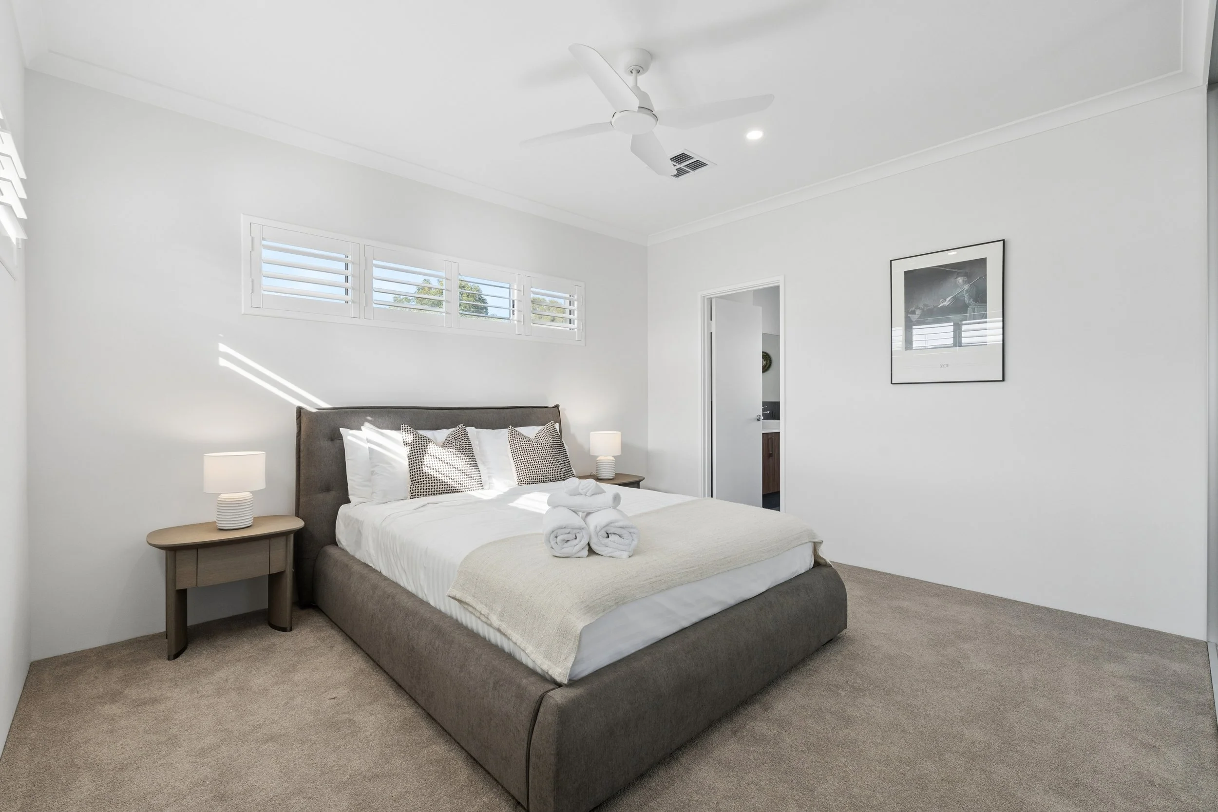 A bedroom with a gray upholstered bed, white bedding, and rolled towels on top. There are two matching bedside tables with white lamps, a framed black and white photo on the wall, and a window with shutters allowing natural light. The room has a ceil