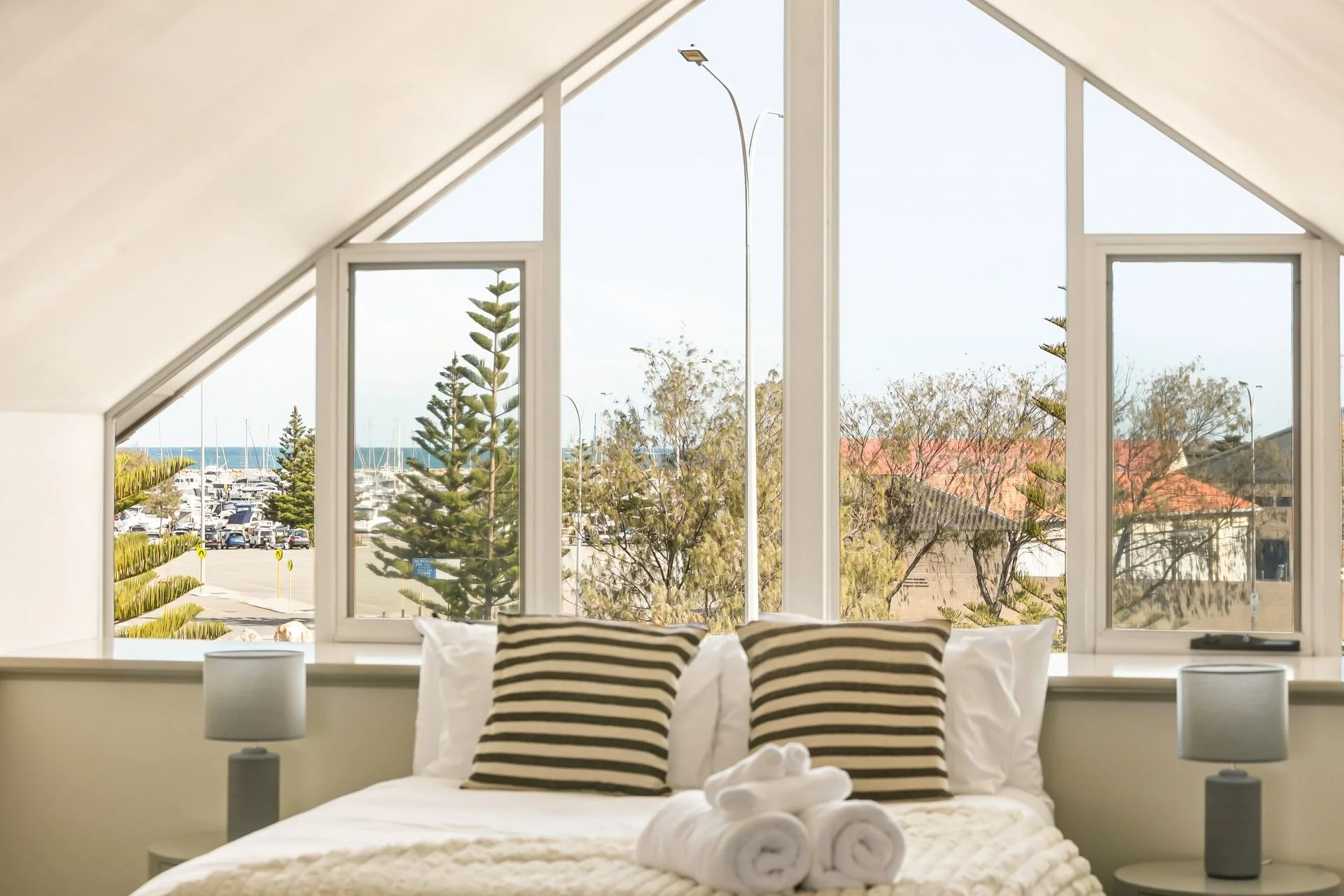 A cozy bedroom with a large arched window showing a view of trees, a parking lot with cars, and a marina in the distance. The bed has white linens, black and white striped pillows, and folded white towels on top. There are two white bedside lamps, on