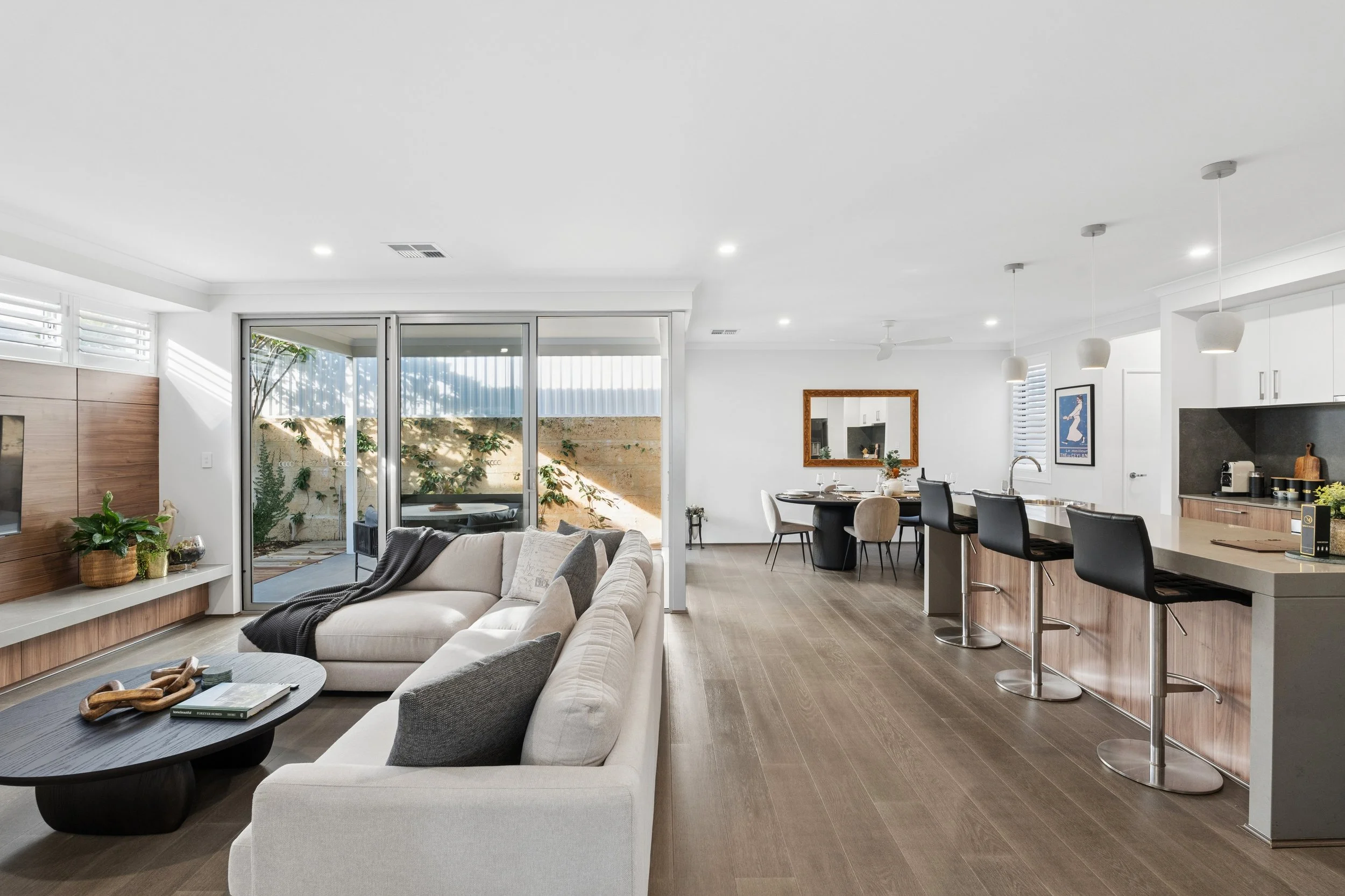 Modern open-concept living room and kitchen with white walls, wooden flooring, sliding glass doors leading outside, a beige sofa, a black coffee table, dining table with chairs, kitchen island with barstools, white cabinets, and minimal decor.