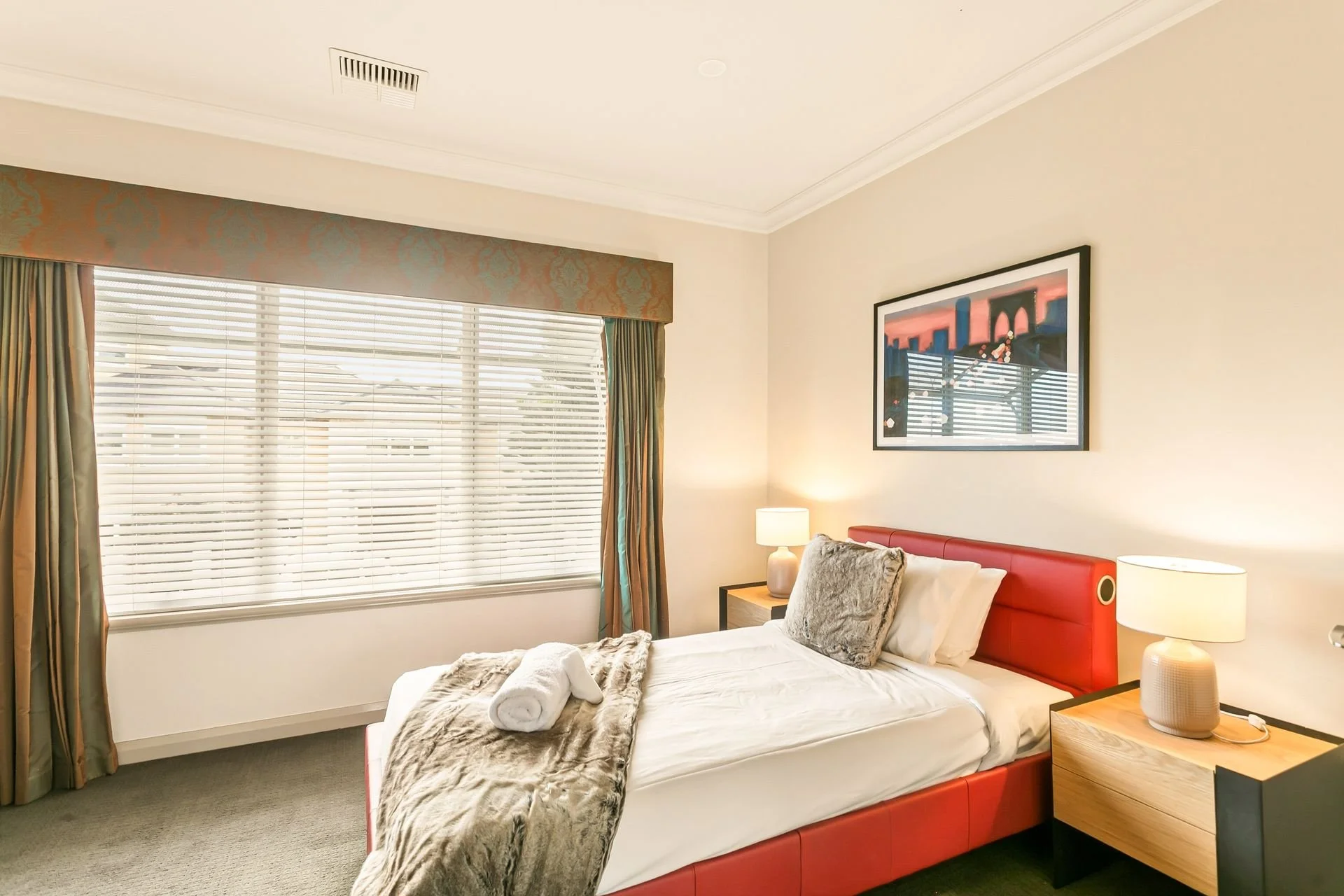 A bedroom with a large window with blinds and curtains, a bed with pillows and a fur throw, bedside tables with lamps, and a framed artwork on the wall.