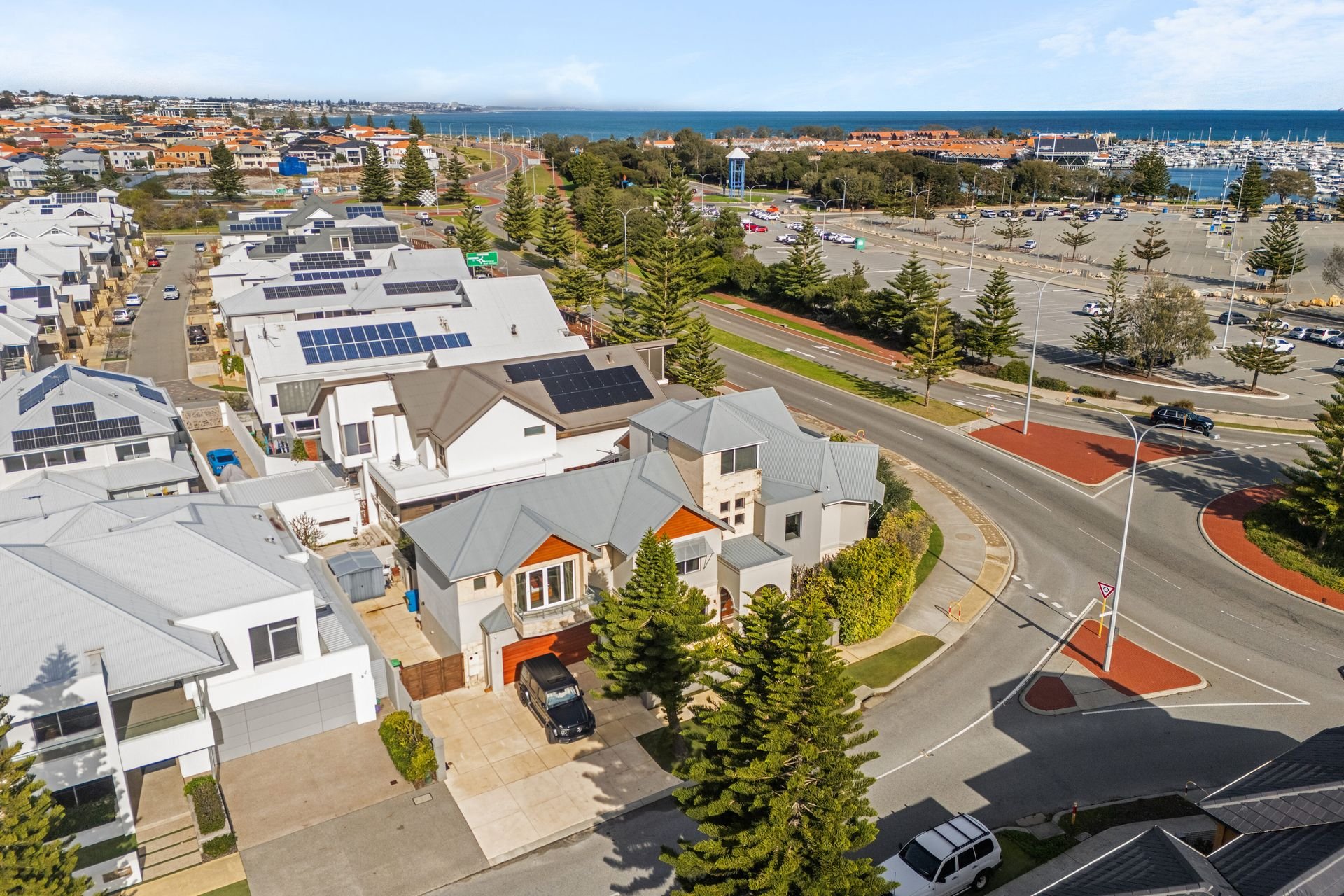 Aerial view of a coastal neighborhood with modern houses equipped with solar panels on rooftops, a street with trees, a parking lot, and a view of the ocean in the background.