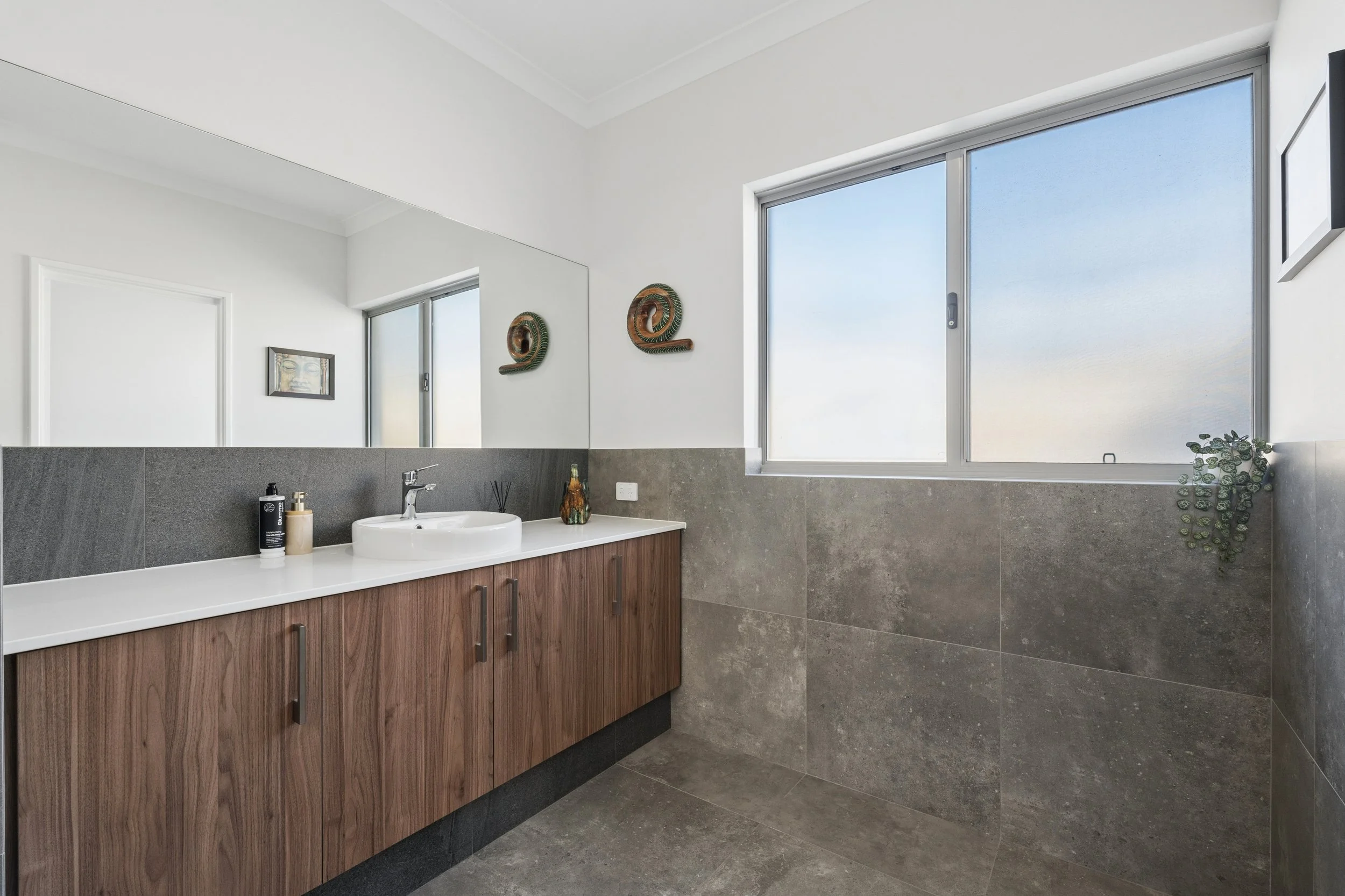 A modern bathroom with white walls, a large window, a sink with a wooden cabinet, decorative wall art, and green plants.