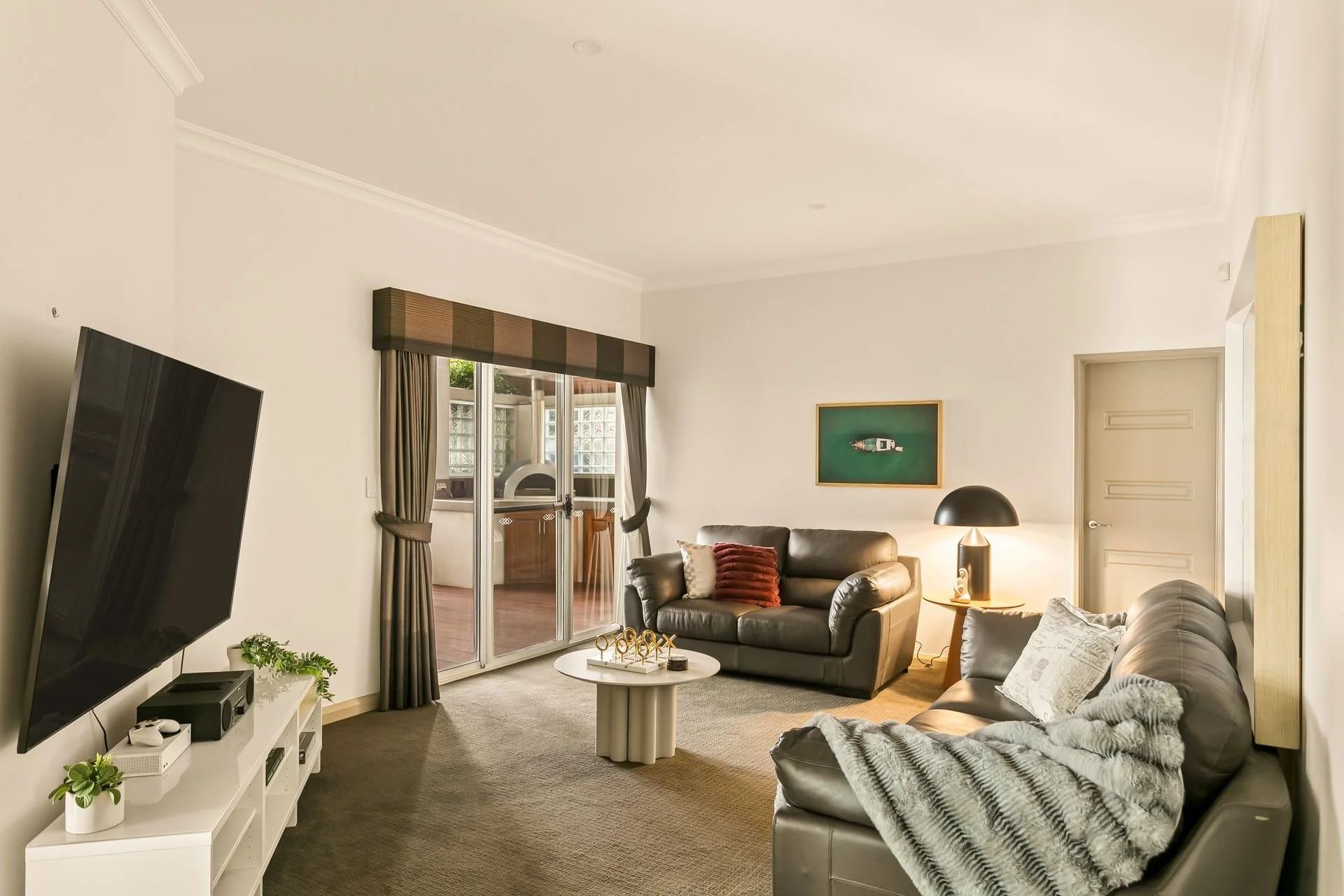 Living room with beige walls, gray leather sofas, a white entertainment unit with plants and electronics, a flat-screen TV, and sliding glass door leading to a balcony.
