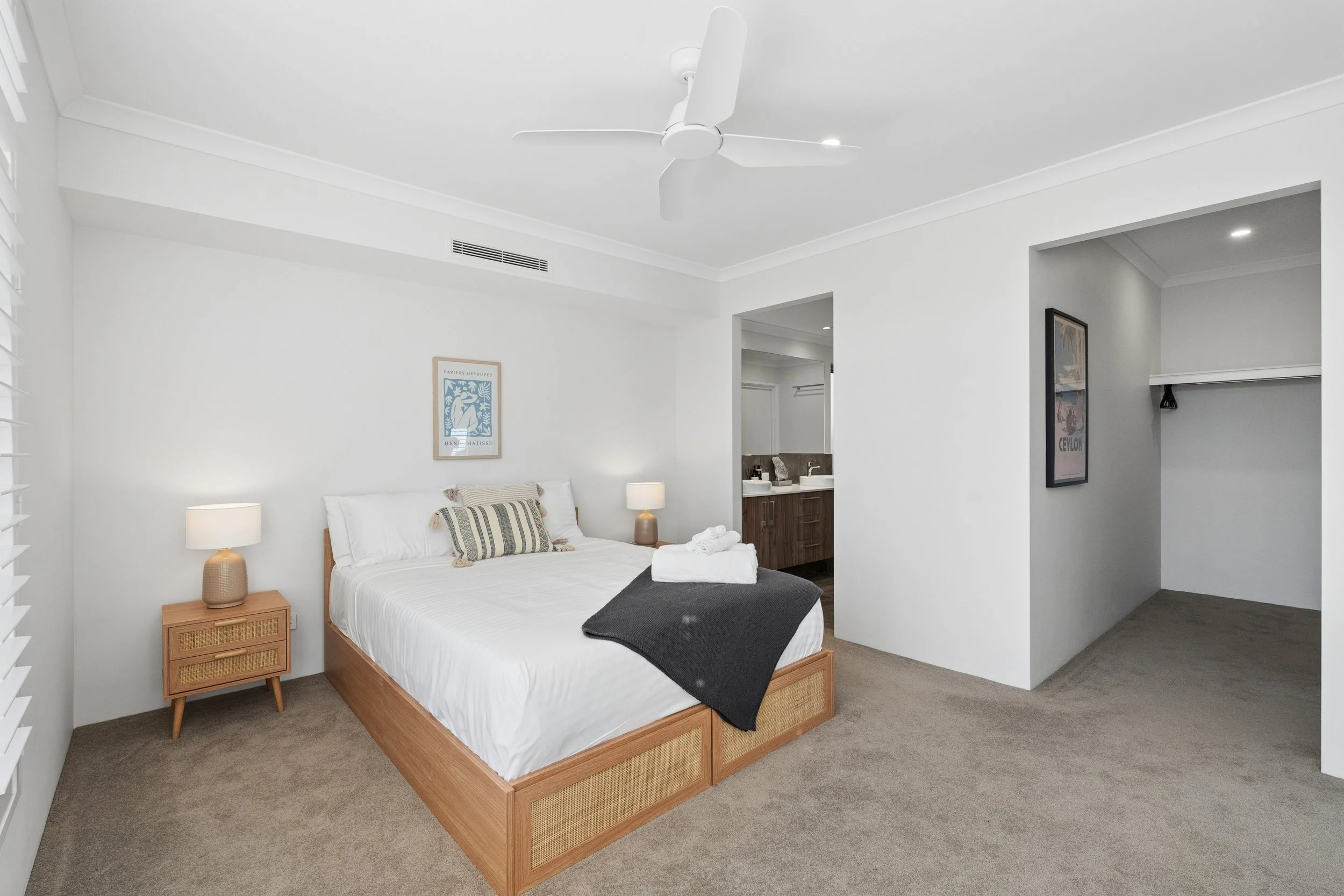 A bedroom with a white bed and wooden frame, two matching nightstands with lamps, a framed art piece on the wall, and a partial view of an adjacent bathroom with a wooden vanity and double sinks.