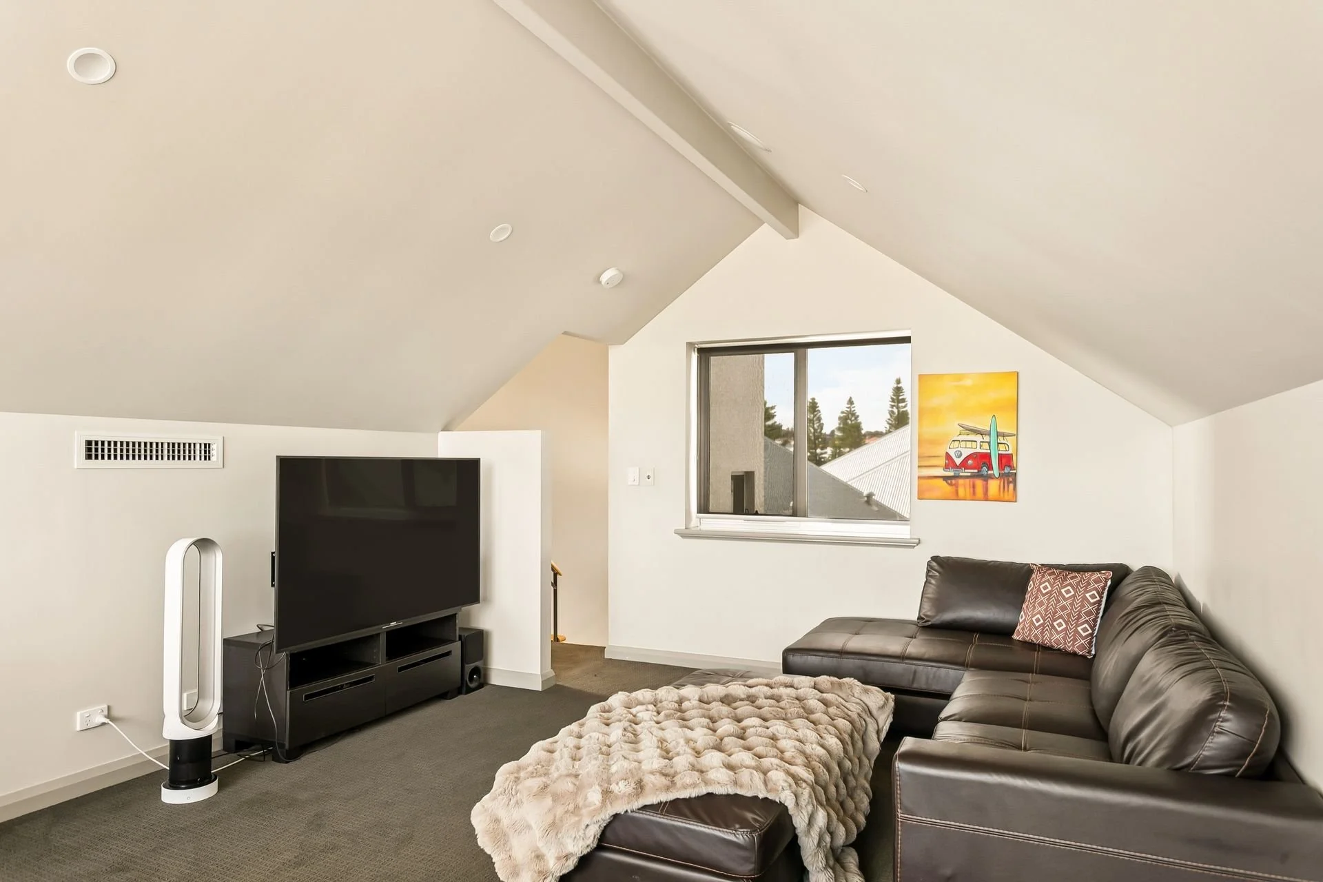 L-shaped living room with a brown leather sofa, a window overlooking trees, a colorful painting of a red van with surfboards, a flat-screen TV on a black stand, a tall white fan, and a beige fuzzy blanket on the sofa.