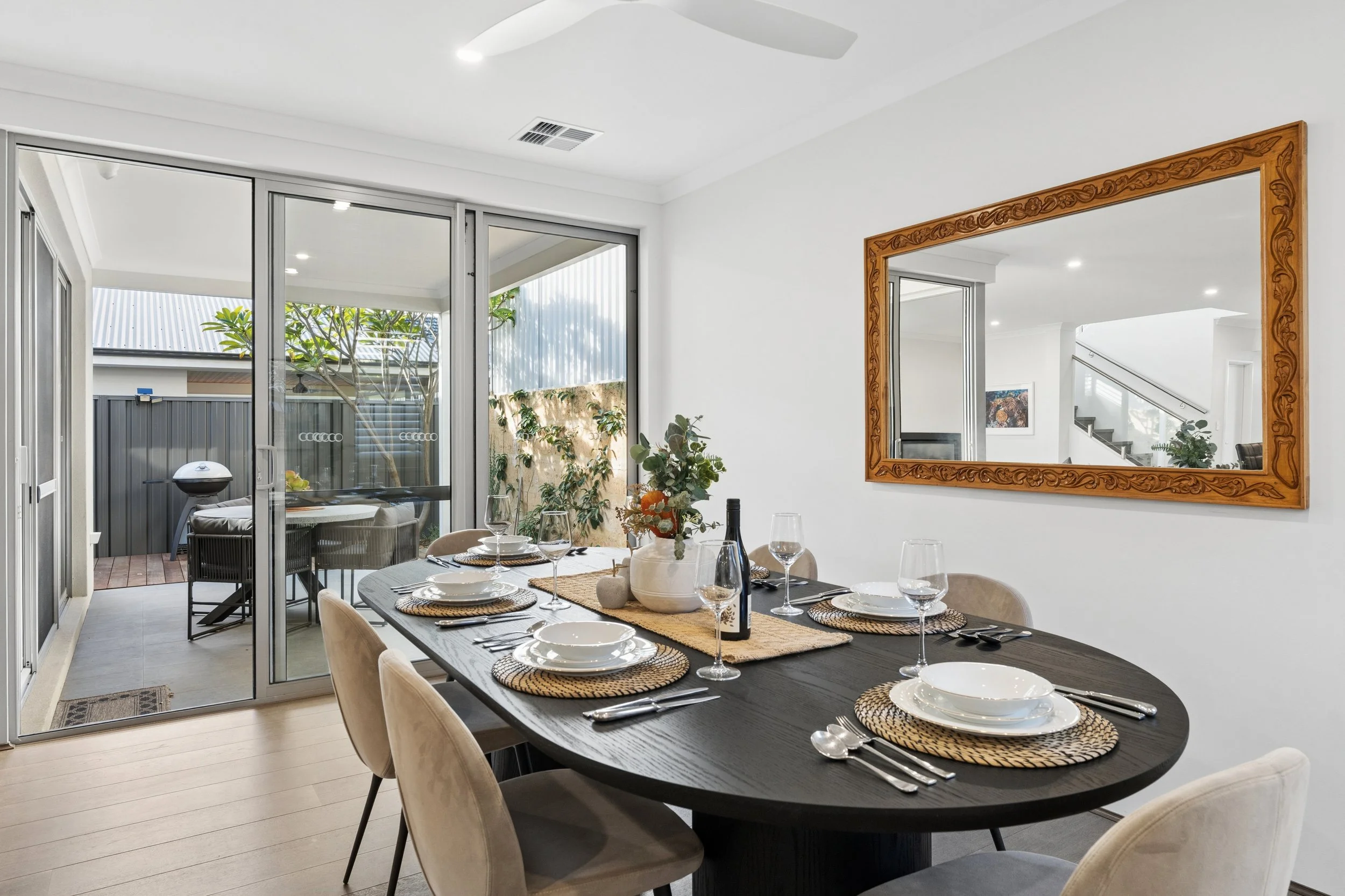 A dining room with a black rectangular table set for a meal with white plates, wine glasses, and woven placemats. Beige upholstered chairs surround the table. A large framed mirror with intricate wood carving hangs on the white wall. Large sliding gl