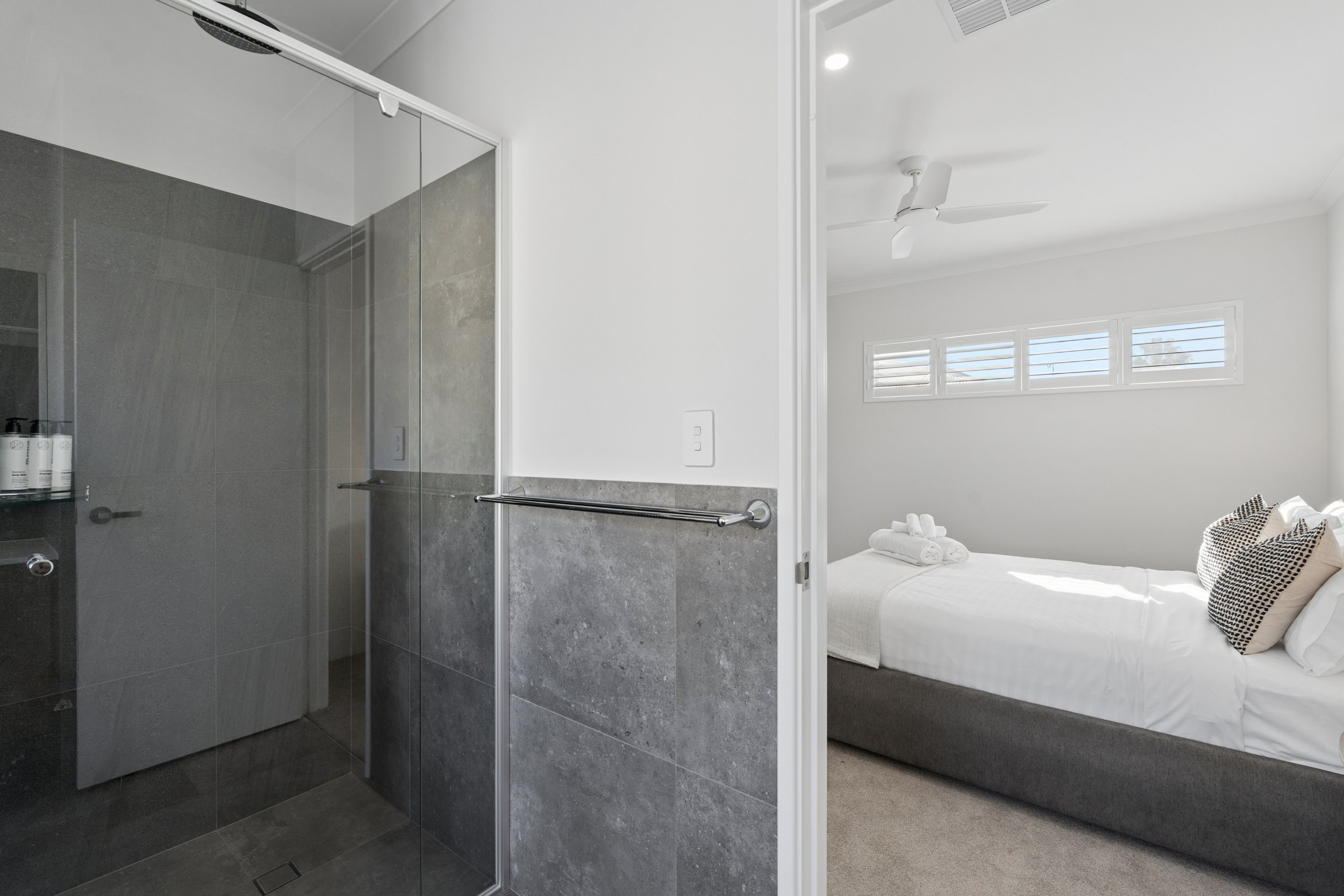 A bathroom with a glass shower enclosure and a bedroom with a white bed and windows.