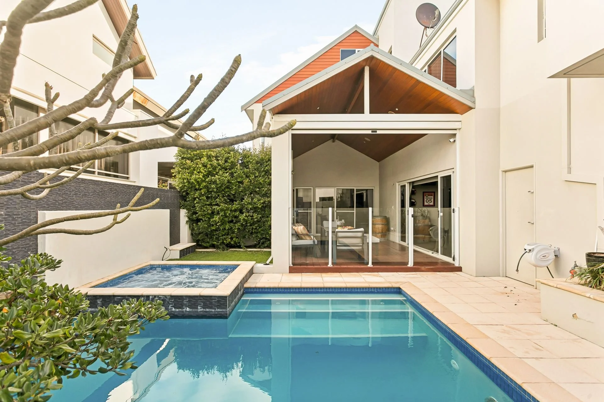 Modern house with a backyard pool, covered patio area with glass sliding doors, surrounded by greenery and neighboring houses.