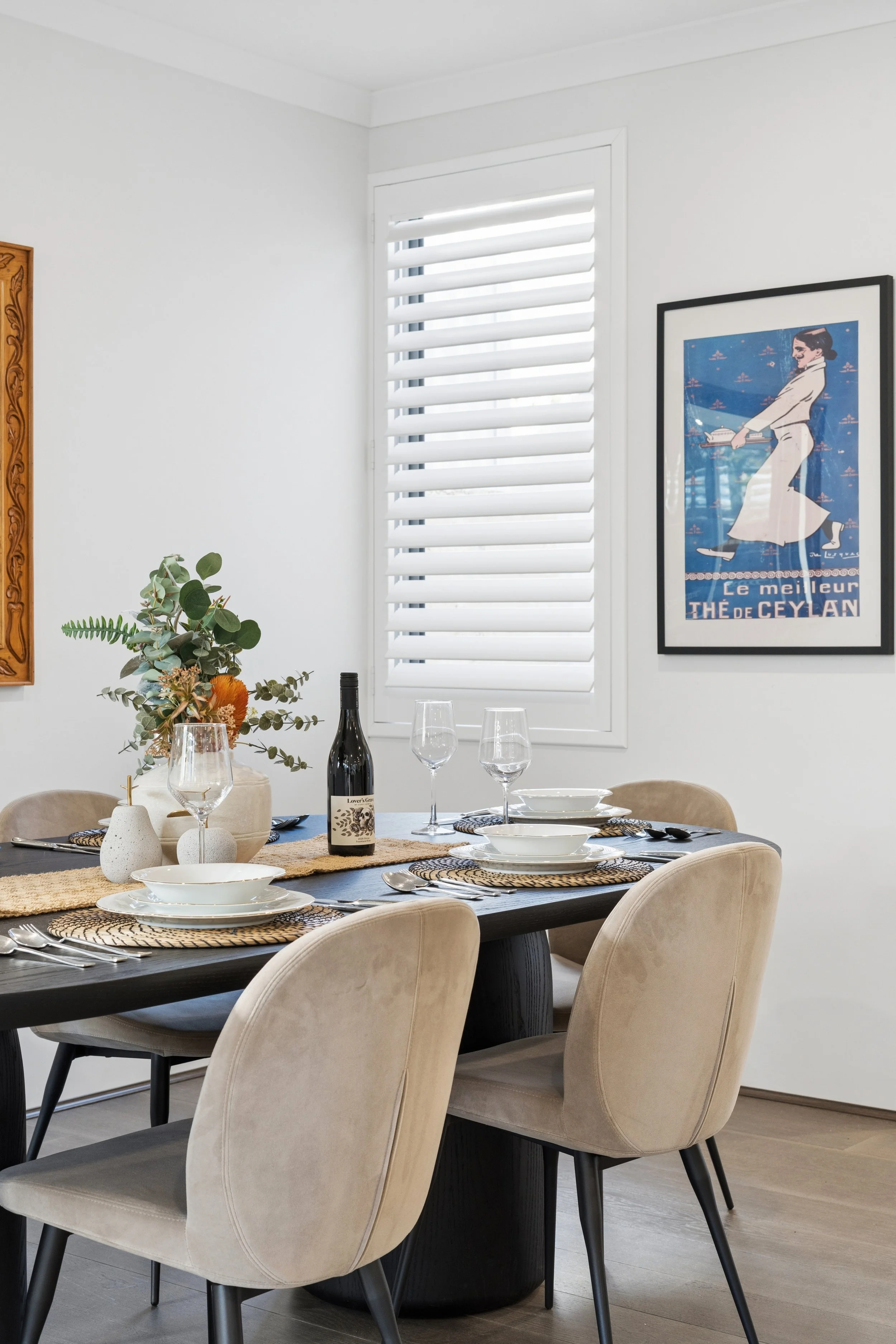 A modern dining room with a black table set with white plates, wine glasses, and cutlery. Beige upholstered chairs surround the table, which has a natural fiber runner. In the background, a window with white plantation shutters, a framed poster, and 