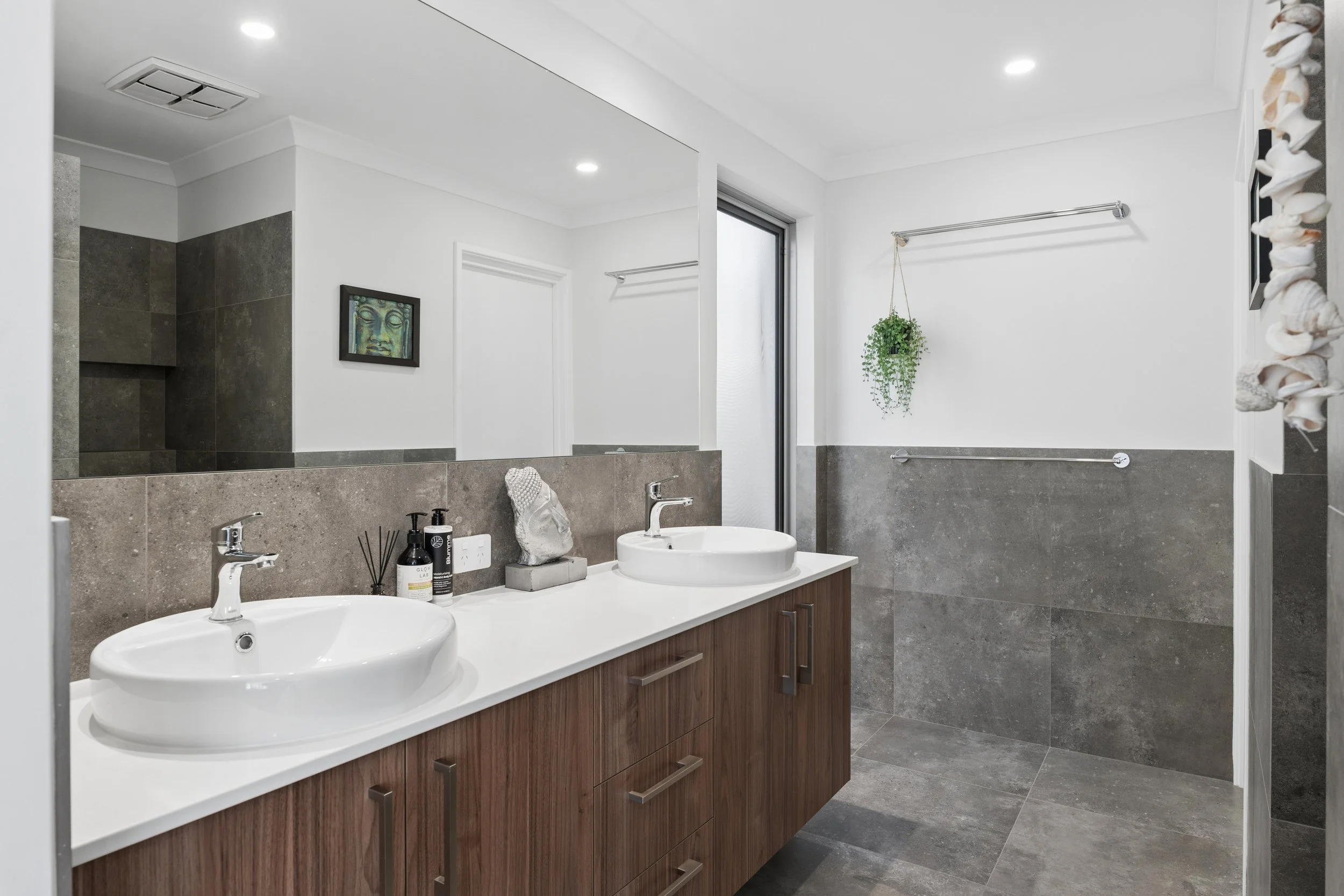 A modern bathroom with double sinks, a large mirror, and gray tiled walls. There is a small decorative statue between the sinks, and a few toiletries on the counter. A window and a hanging plant are visible, along with towel bars on the wall.