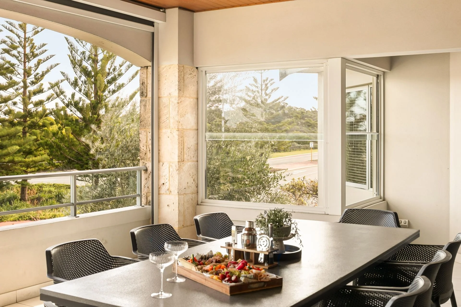 Indoor dining area with a large rectangular table, black mesh chairs, and a tray of assorted finger foods and desserts. Large windows with views of trees and a street outside, decorated with white walls and a stone accent wall.