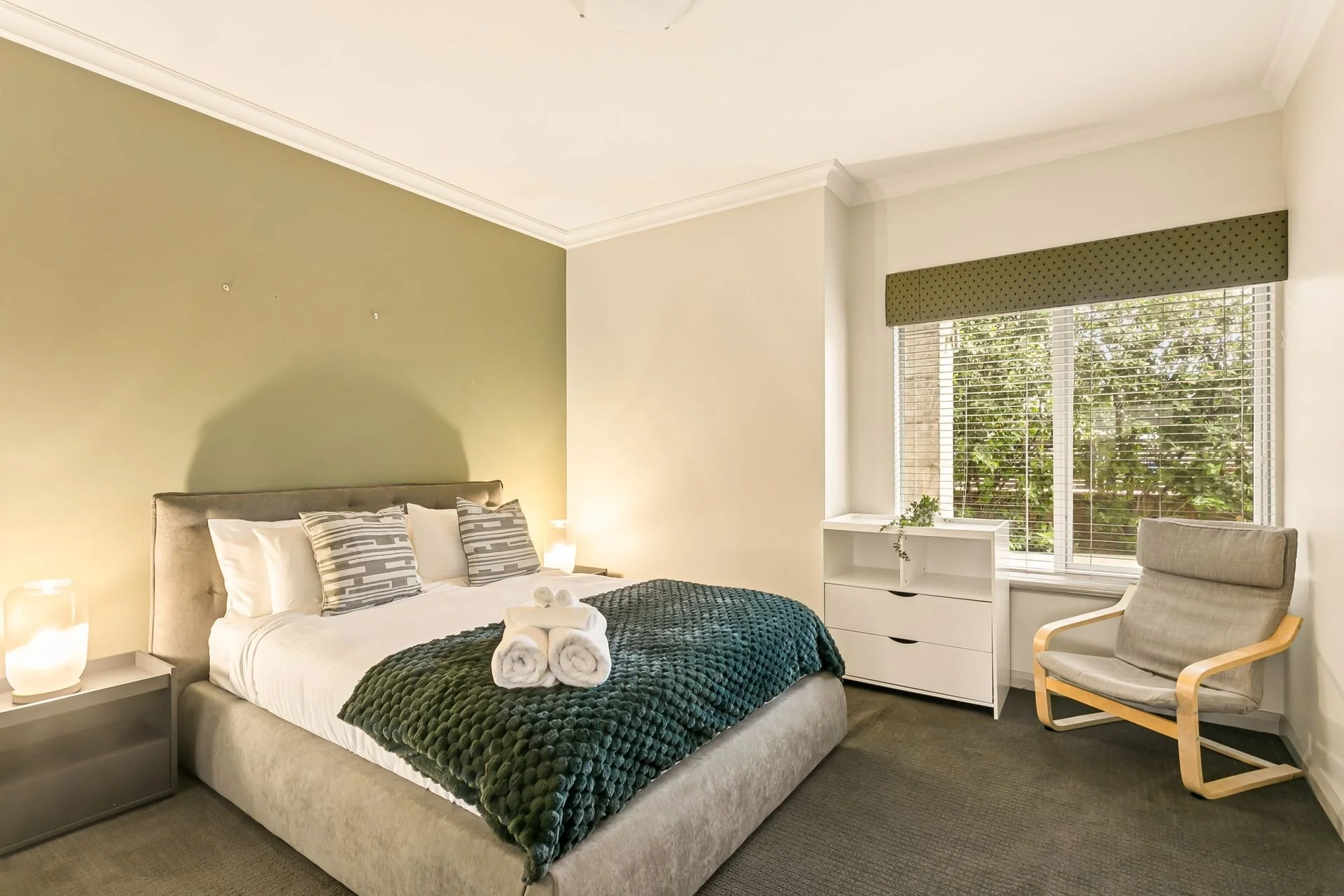 Bright bedroom with a gray upholstered bed, green textured blanket, white sheets, and decorative pillows; nightstand with a lamp; white dresser with a small plant; large window with blinds and green valance; gray armchair with wooden frame.