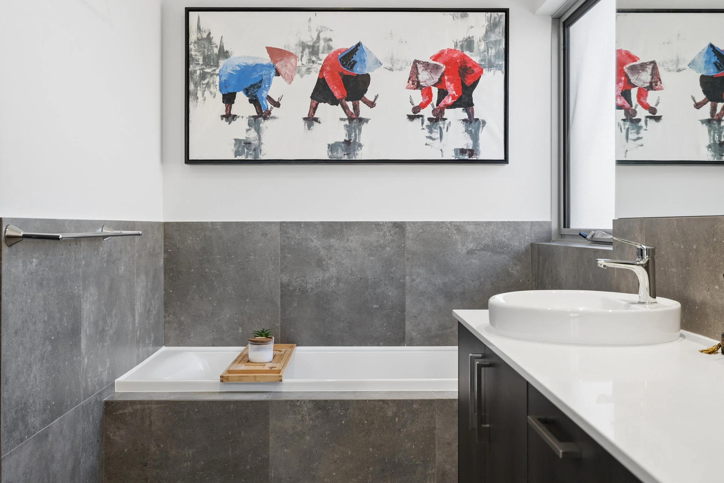Modern bathroom with gray tile walls, a white bathtub with a wooden bath tray holding a candle, a small potted plant, a white sink, and a large colorful abstract painting of people in traditional Asian conical hats on the wall.