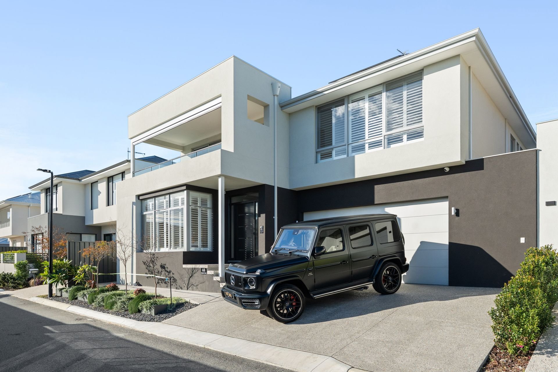 Modern multi-story house with white and black exterior, large windows with blinds, and a black Mercedes-Benz G-Class parked in the driveway. Well-maintained landscaping with small trees and shrubs in front.