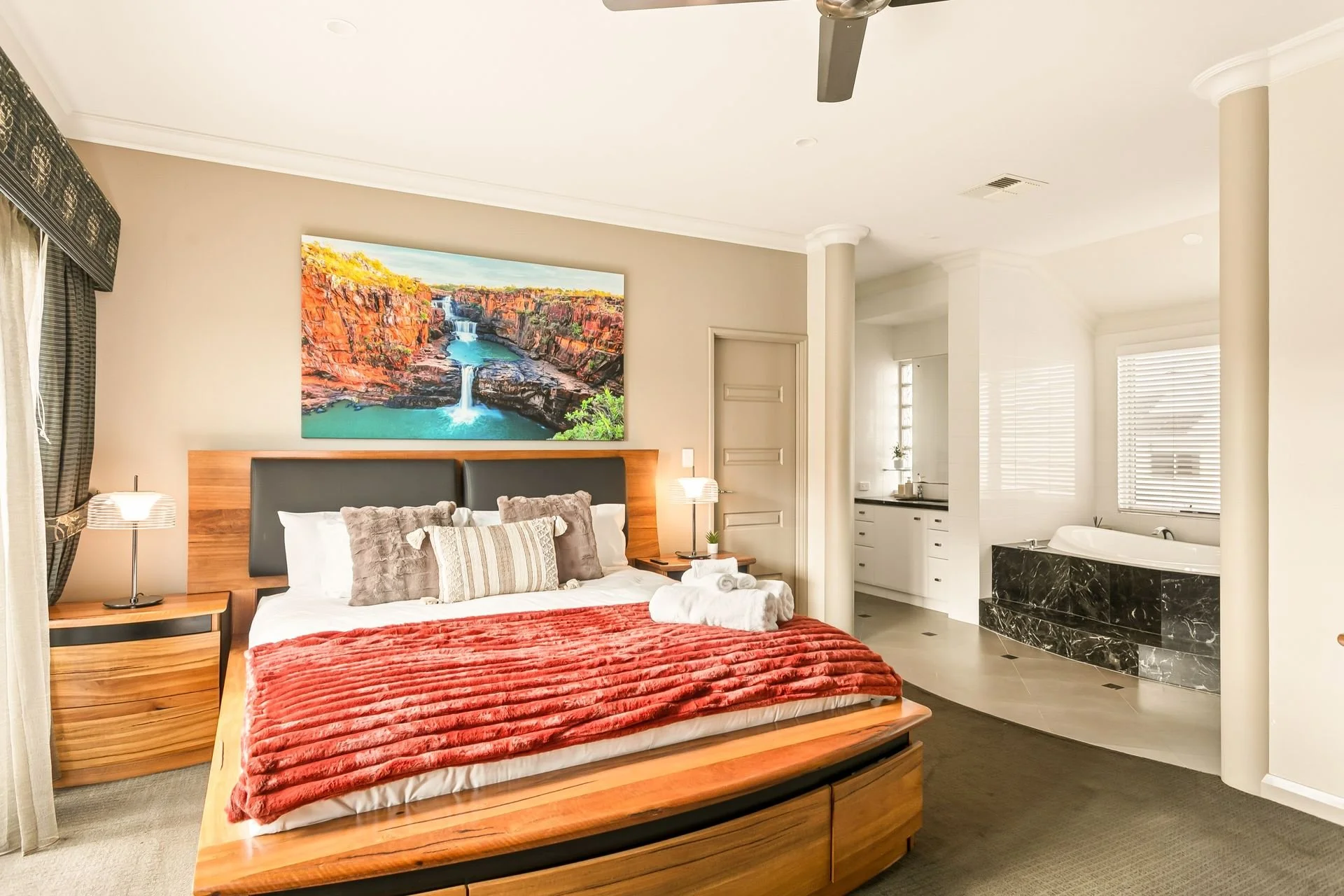Modern bedroom with wooden bed frame, red velvet blanket, gray and striped pillows, nightstand with lamp, large painting of a waterfall on the wall, and an open bathroom with a bathtub and window blinds.