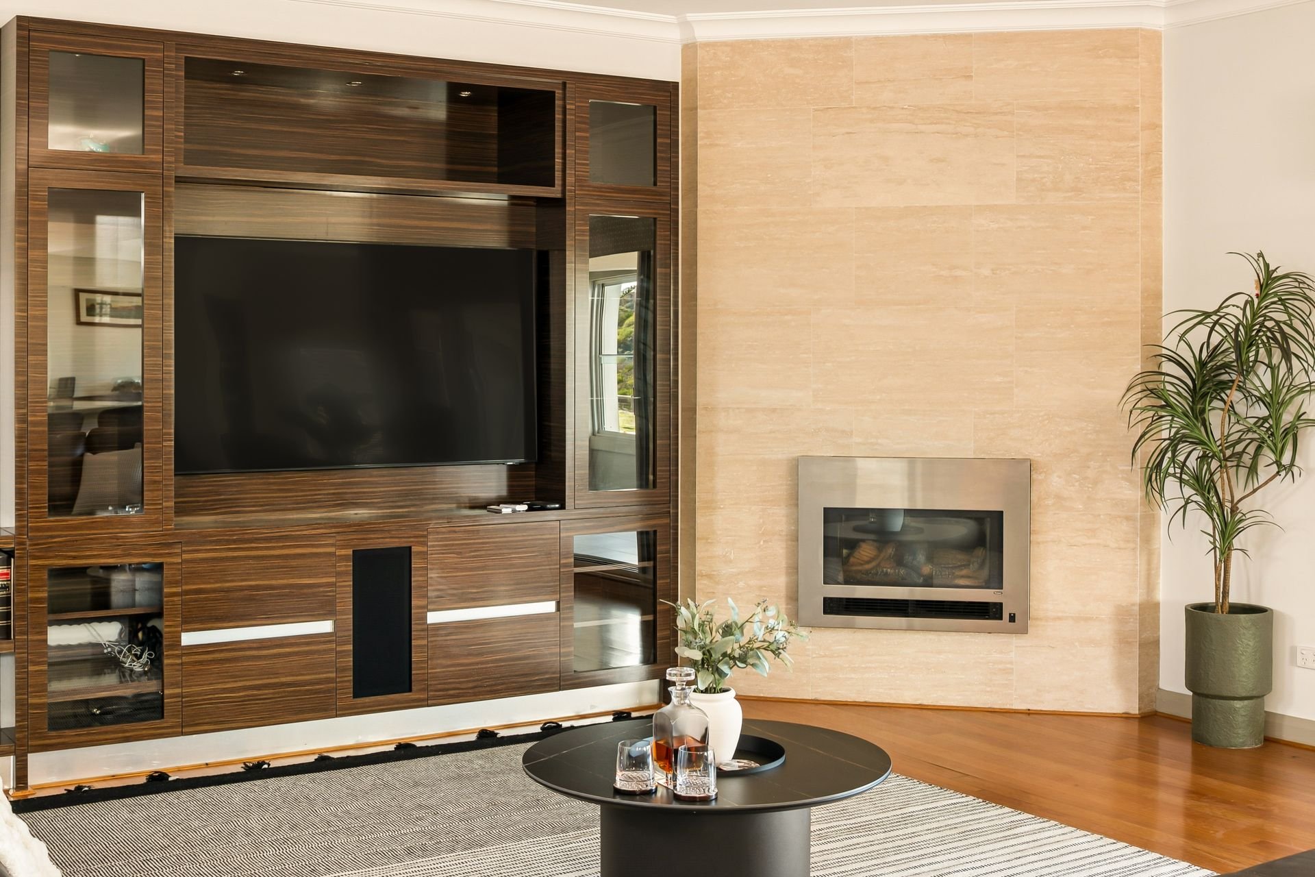 Living room with a built-in wooden entertainment center surrounding a large flat-screen TV. To the right, a modern fireplace is embedded in a beige stone wall. A tall potted plant is placed beside the fireplace. A black round coffee table holds a whi