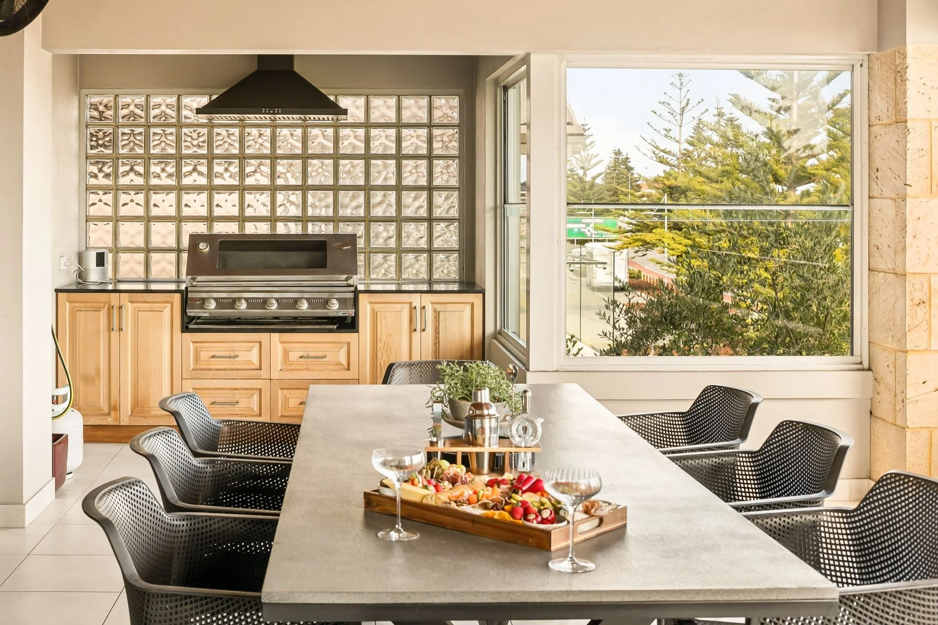 A dining area with a large rectangular table surrounded by black chairs, a serving tray with vegetables, plate, and drinks, a kitchen with a glass block wall behind a stove, and large windows showing trees and a parking lot outside.