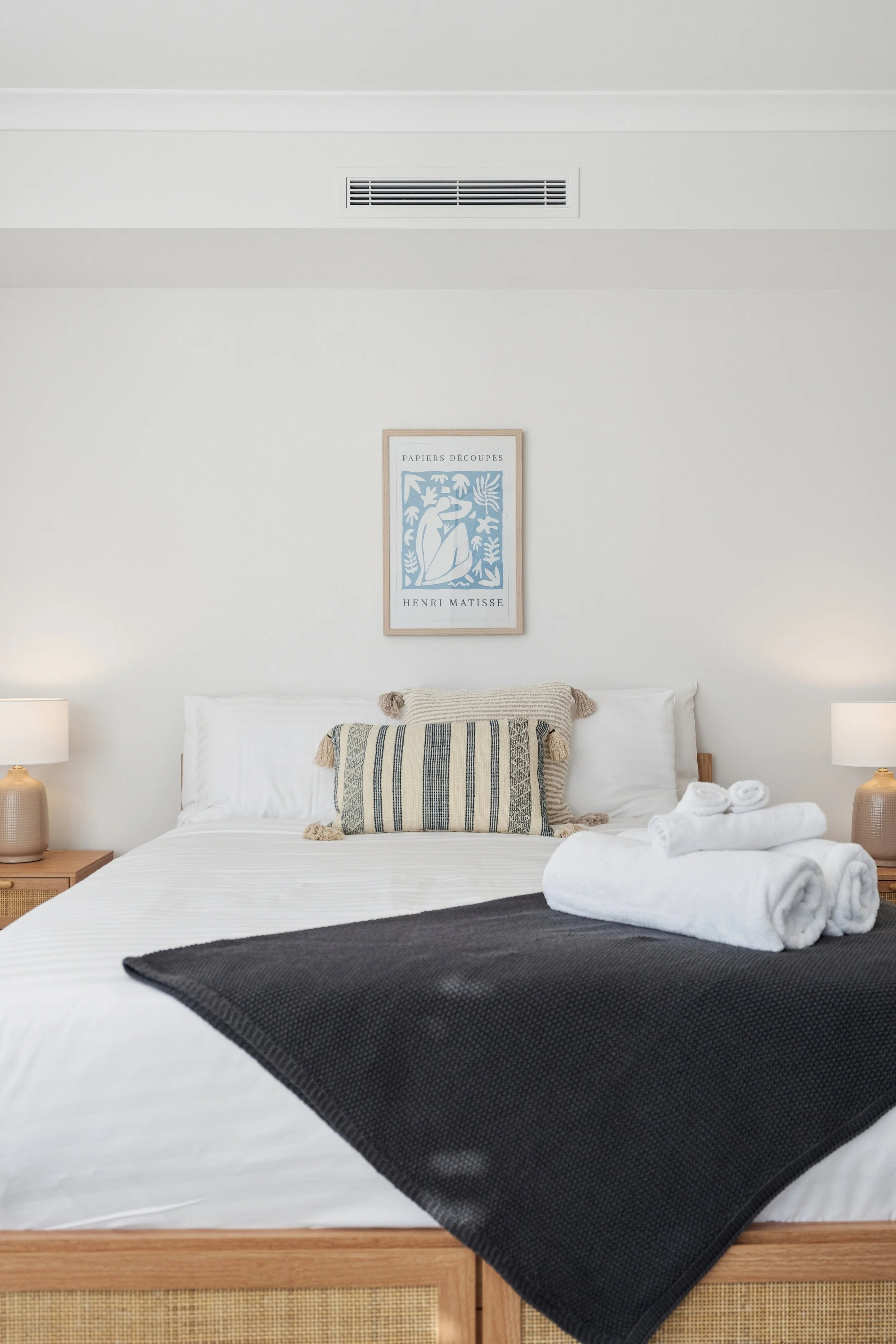 A neatly made bed with white bedding, a black throw blanket, and a decorative pillow with striped pattern. Rolled towels are placed at the foot of the bed. There are two table lamps on either side of the bed, and a framed Henri Matisse artwork hangs 