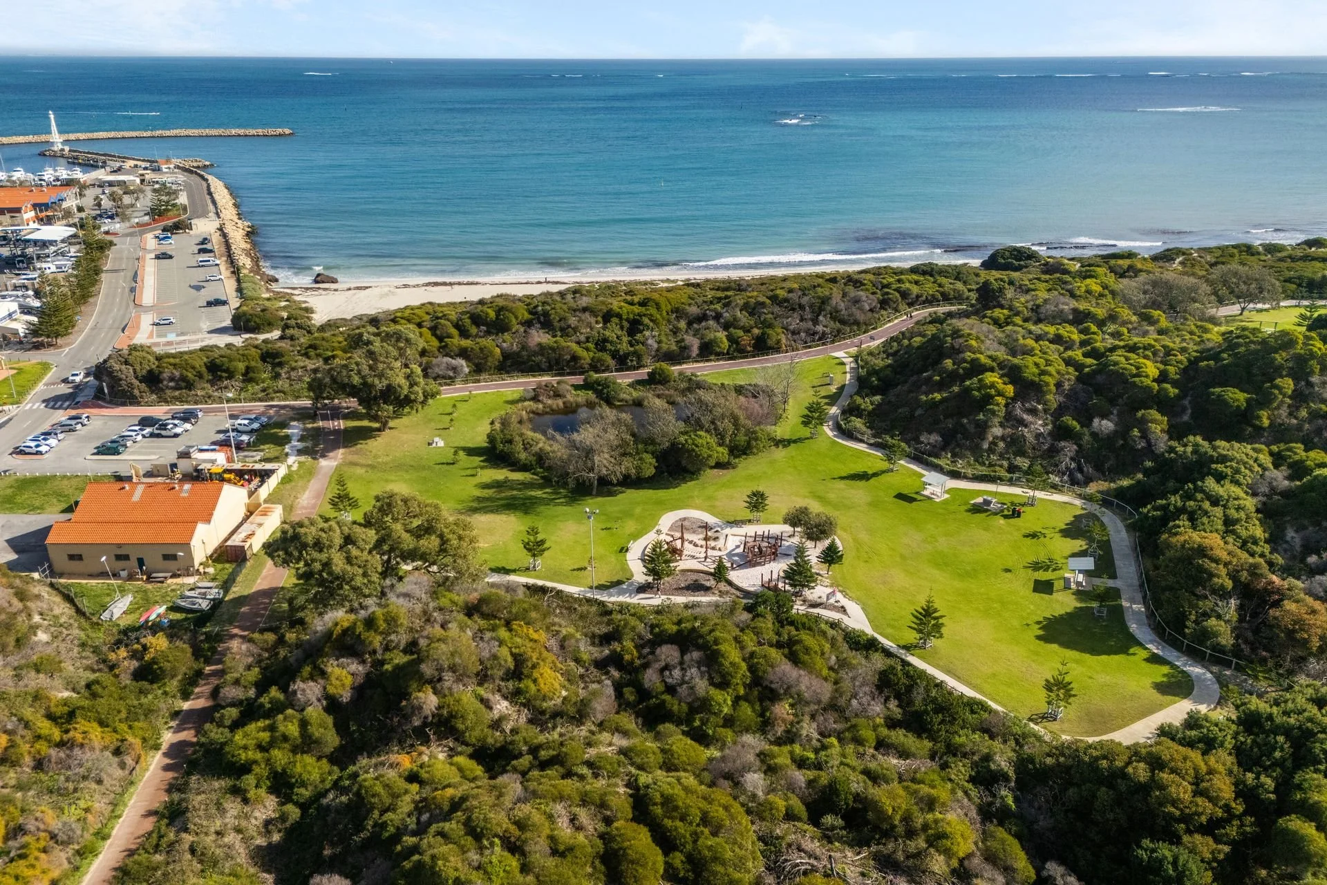 Aerial view of a coastal park with lush greenery, walking paths, playground equipment, overlooking a beach and ocean with a breakwater and harbor.