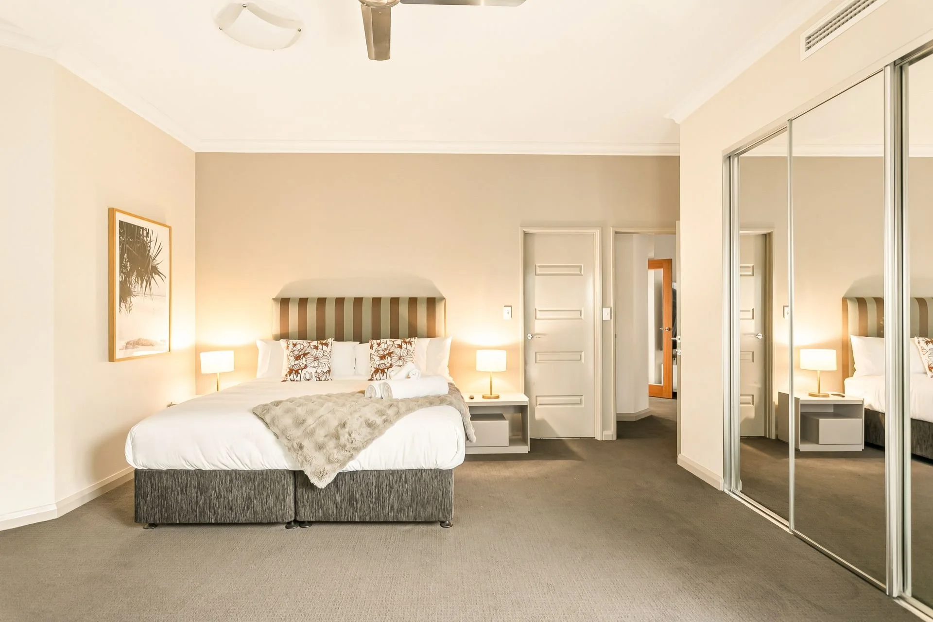 A neatly made bedroom with a large bed featuring a striped headboard, topped with white pillows and floral decorative pillows. Flanking the bed are two bedside tables, each with a lit white lamp. The room has beige walls, a piece of framed wall art o