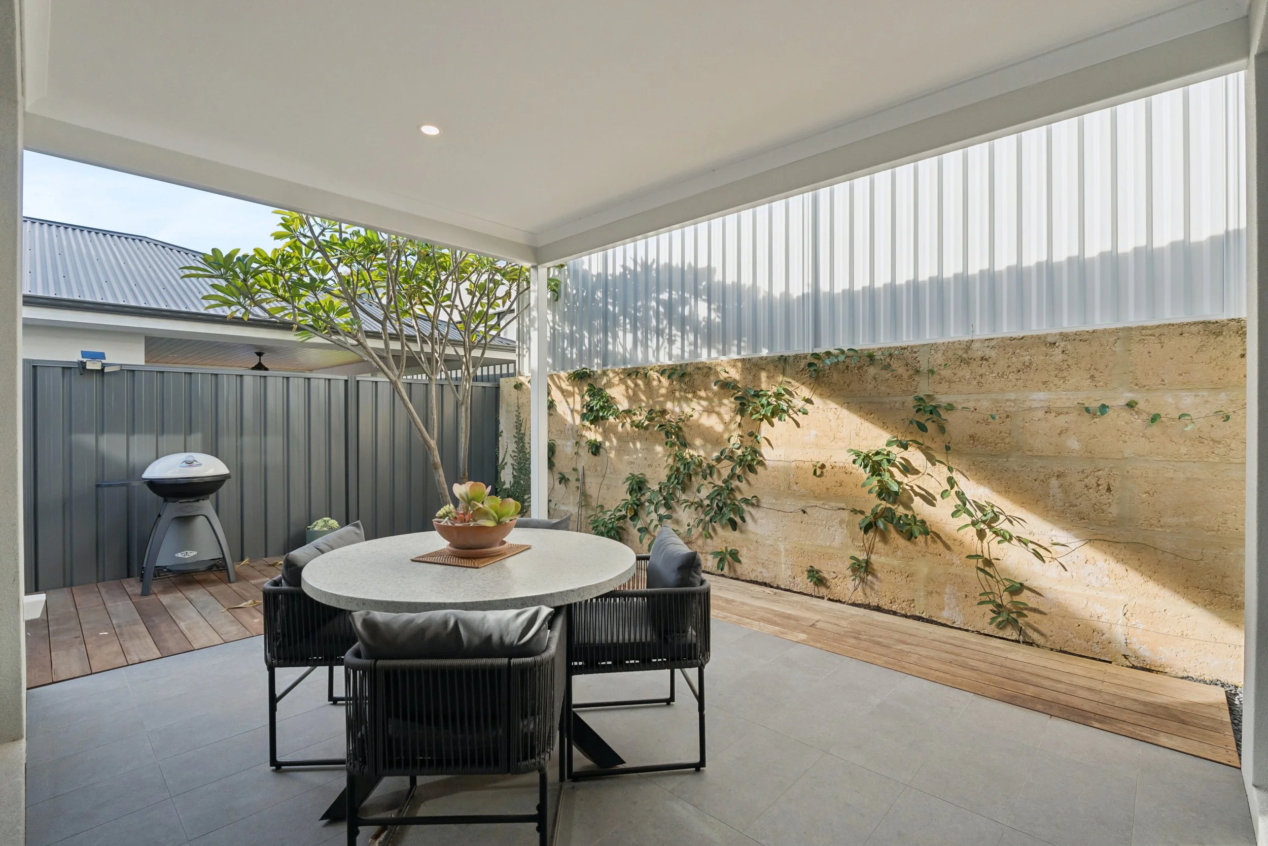 Covered outdoor patio with a round table, six chairs, a potted plant, and a barbecue grill, enclosed by a wooden sliding door and beige brick wall with vines.
