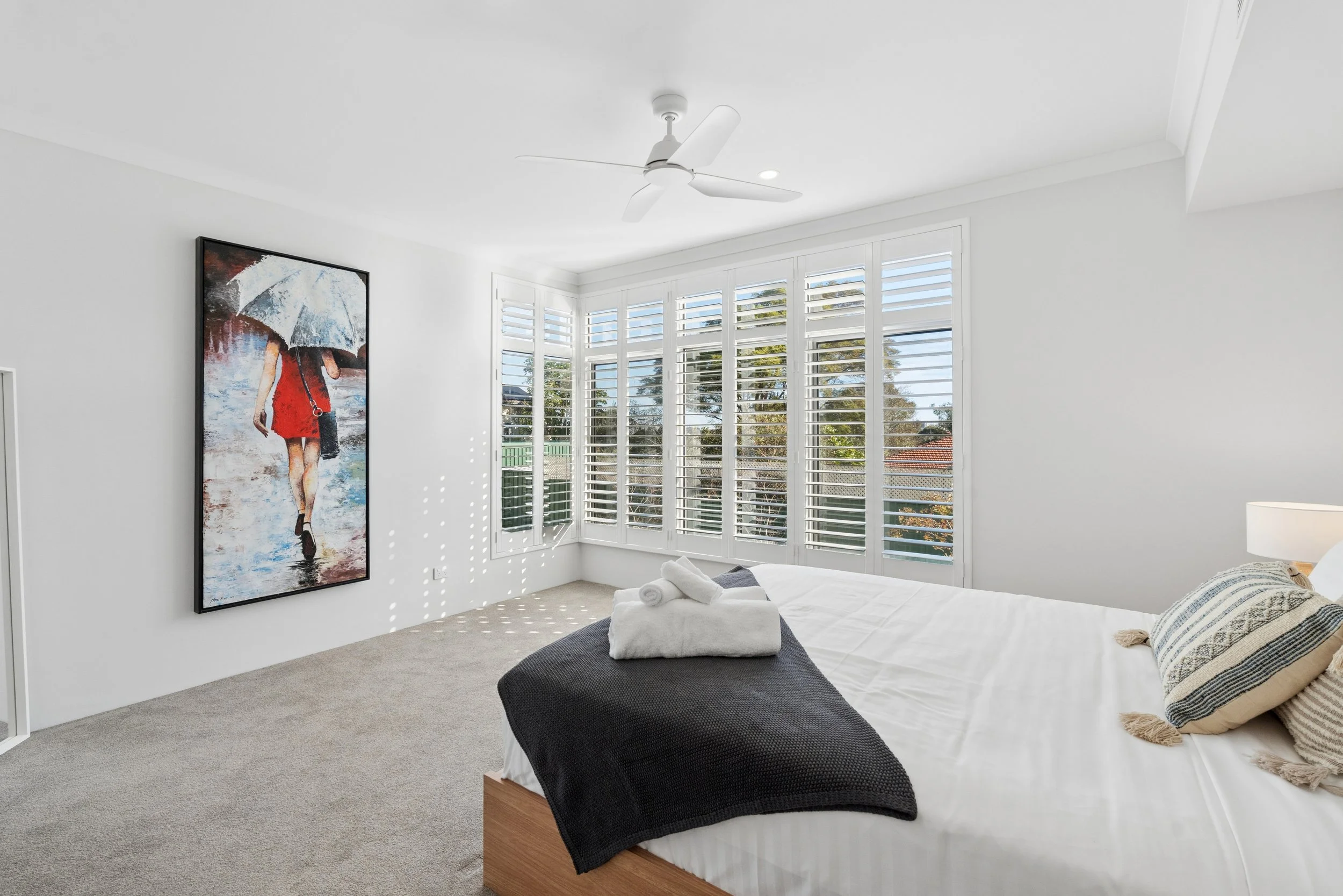 Bright bedroom with large window shutters, a bed with white linens and a black blanket, and a modern ceiling fan.
