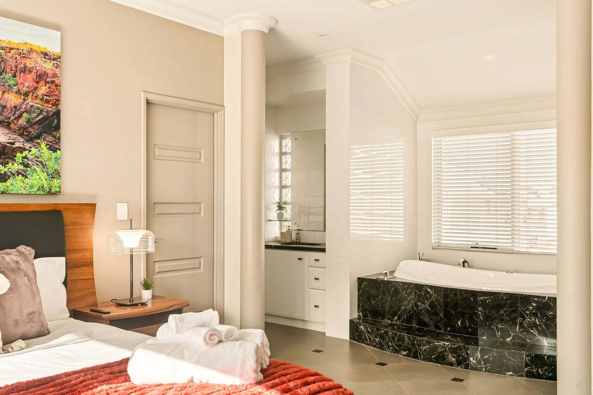 A cozy bedroom with a bed that has white linens, pillows, and a burnt orange blanket. A small wooden side table next to the bed holds a modern white lamp and a small potted plant. Open to a bathroom with a large bathtub surrounded by black marble, wh