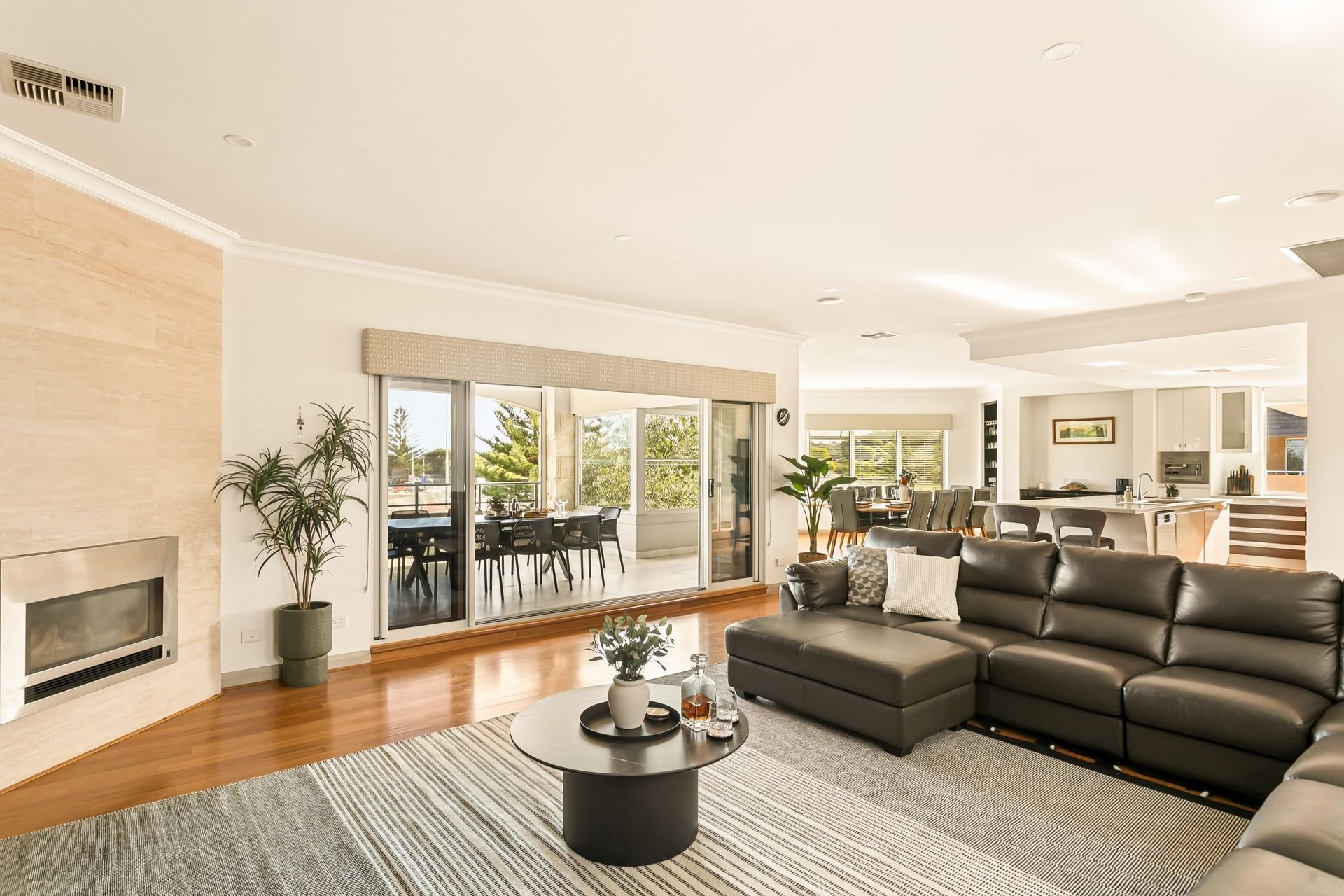 Modern living room with a black leather sectional sofa, a round coffee table with decor items, and large windows with natural light, leading to an outdoor dining area with a table and chairs.