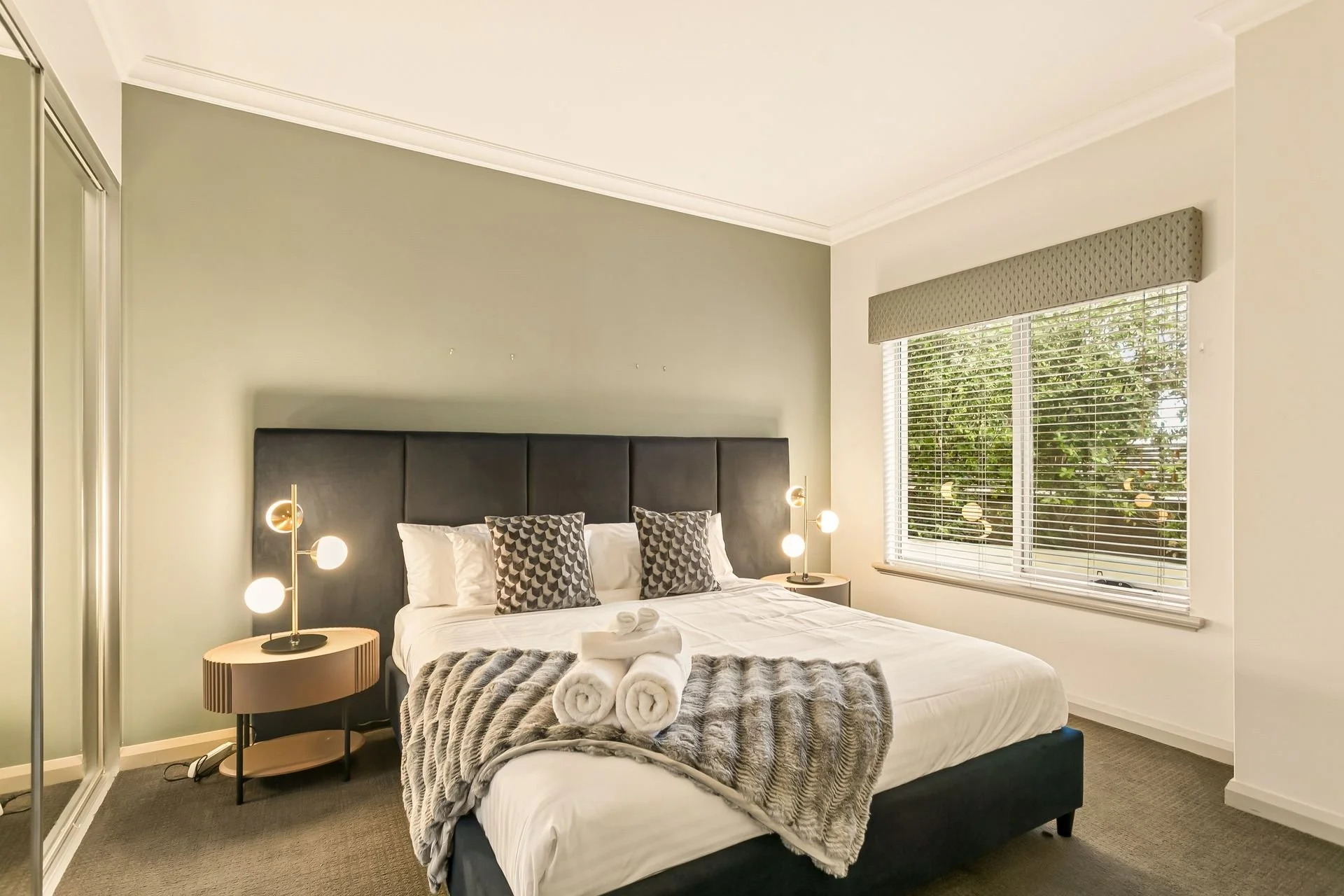 A neatly made bed with white sheets, decorated with patterned pillows, a faux fur blanket, and towels shaped into swans. Two modern bedside lamps with round bulbs sit on bedside tables, and a large window with blinds and a valance shows greenery outs